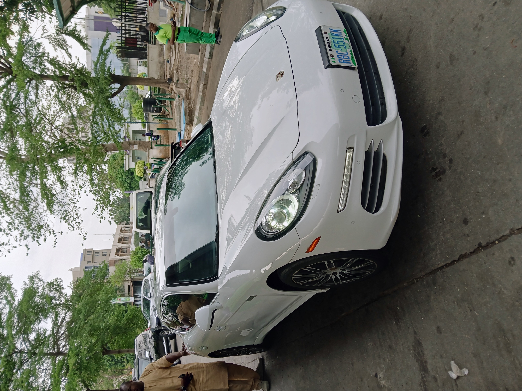 A white Porsche Panamera is parked on the side of a street. The license plate reads "ABUJA RBC-551 KK". A man in a tan traditional garment stands next to the car. In the background, there are other parked vehicles, trees, buildings, and individuals in green uniforms performing what appears to be road maintenance. The location is within the city of Abuja, Nigeria.