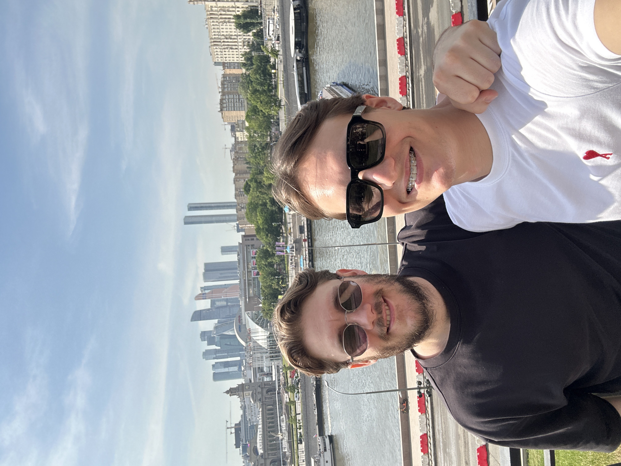 Two adult males are depicted posing for a selfie on a bright, sunny day in Moscow, Russia. The man on the left has light brown hair, a full beard, and wears round, tinted sunglasses and a plain black crew-neck t-shirt. He is smiling. The man on the right, with light brown hair, is clean-shaven and wears dark, rectangular sunglasses, a white crew-neck t-shirt with a small red heart logo, and dental braces visible on his upper teeth. He is smiling broadly and has his left arm casually resting on the left man's right shoulder.
The background reveals a panoramic view of the Moskva River and parts of the city. Distinctly visible on the distant horizon are the numerous modern skyscrapers of Moscow City (Moscow International Business Center). To the left, a large, ornate building with a prominent clock tower and classical architectural elements is identifiable as the Kievsky Railway Station, situated near the river. The Moskva River flows horizontally through the midground, with at least two boats or ferries docked along its far bank. Buildings of varying architectural styles and densities line the riverbanks, interspersed with trees. Construction cranes are visible in several areas. In the immediate foreground, a concrete barrier or wall with red and white striped road barriers is present, along with a grassy patch and parts of a metal railing. The sky is light blue with sparse, wispy clouds, indicating clear weather.