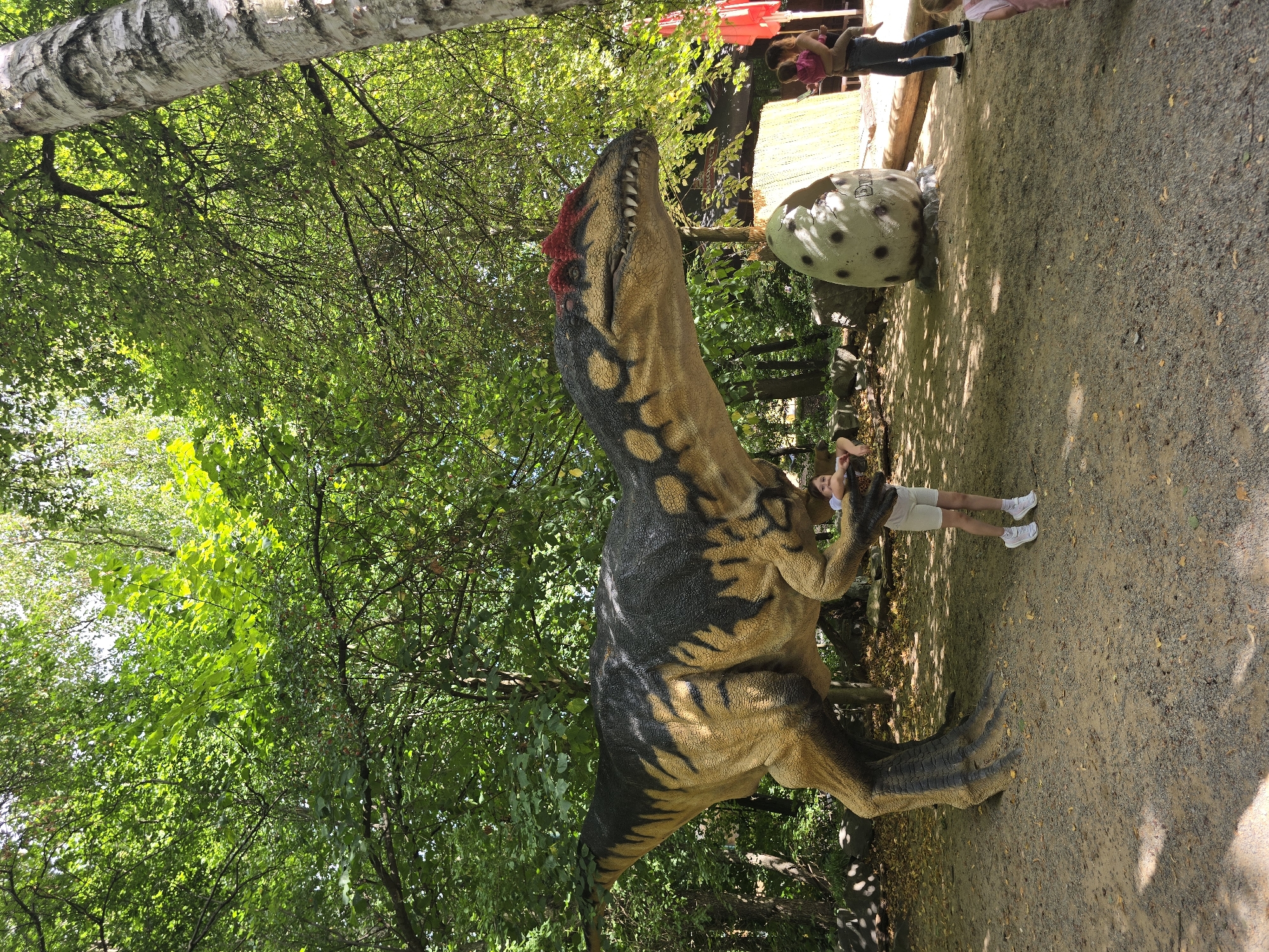 A full-body, eye-level, outdoor shot in daylight shows a large, realistic dinosaur model in a wooded area. The dinosaur model, likely a depiction of a Carnotaurus, has a brown and black patterned body and a red frill around its head, with sharp teeth visible in its open mouth. A young girl in a white t-shirt and light-colored shorts stands on the sandy ground in front of the dinosaur, looking towards it. Behind the dinosaur, a large, broken dinosaur egg model rests on the ground. To the right, a woman in a pink shirt and dark pants holds a young child, and another child in a pink shirt and white shorts is partially visible further to the right. The environment is characterized by dense green foliage, with sunlight filtering through the trees and casting dappled shadows on the ground. The location is identified as the DinoPark Vyškov in the city of Vyškov, Czechia.