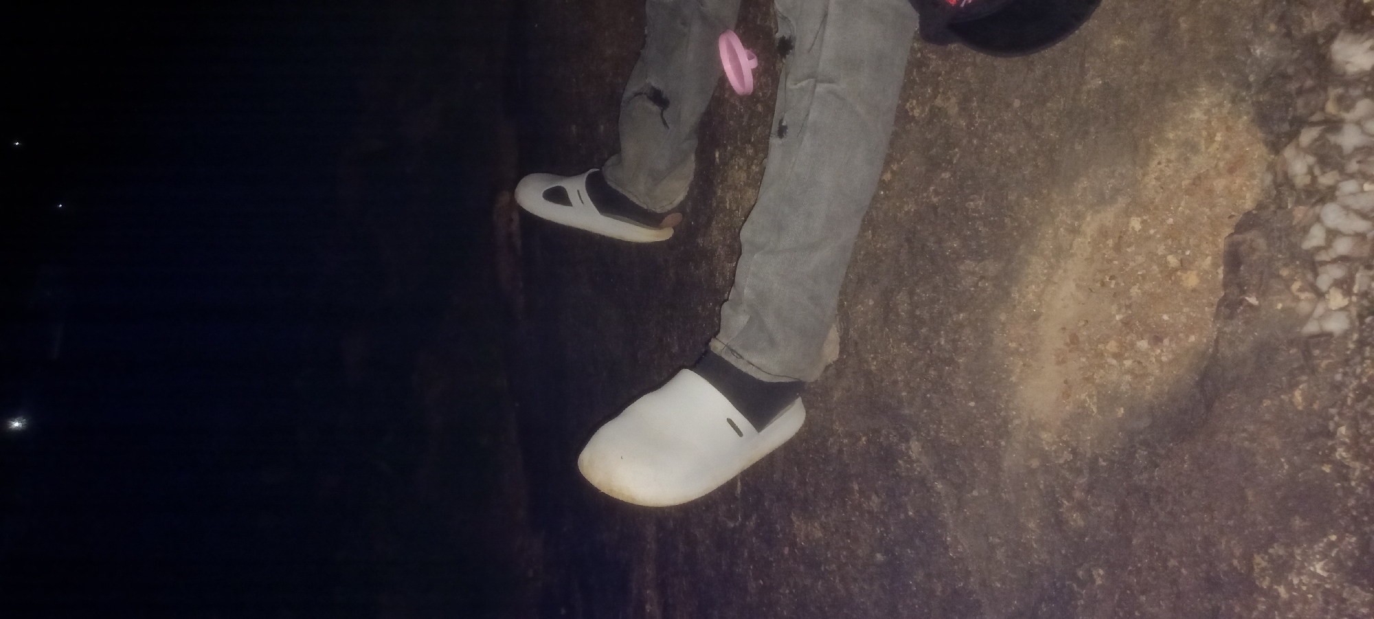 The image shows two individuals lying on the ground at night, their legs extended. The ground appears to be a mix of dirt and gravel. The individuals are wearing casual clothing, including jeans and shoes. One person is wearing white slip-on shoes and black socks, while the other is wearing white slip-on shoes with a black accent. A pink ring-shaped object is visible near the right leg of one individual, and a black object, possibly a bag or hat, is partially visible in the bottom right corner. The background is dark and indistinct, suggesting an outdoor environment at night, possibly in a rural or less developed area, not specifically identifiable as Kwang, Nigeria. There is no indication of any specific activity or interaction between the individuals, other than them being in a resting or reclined position.