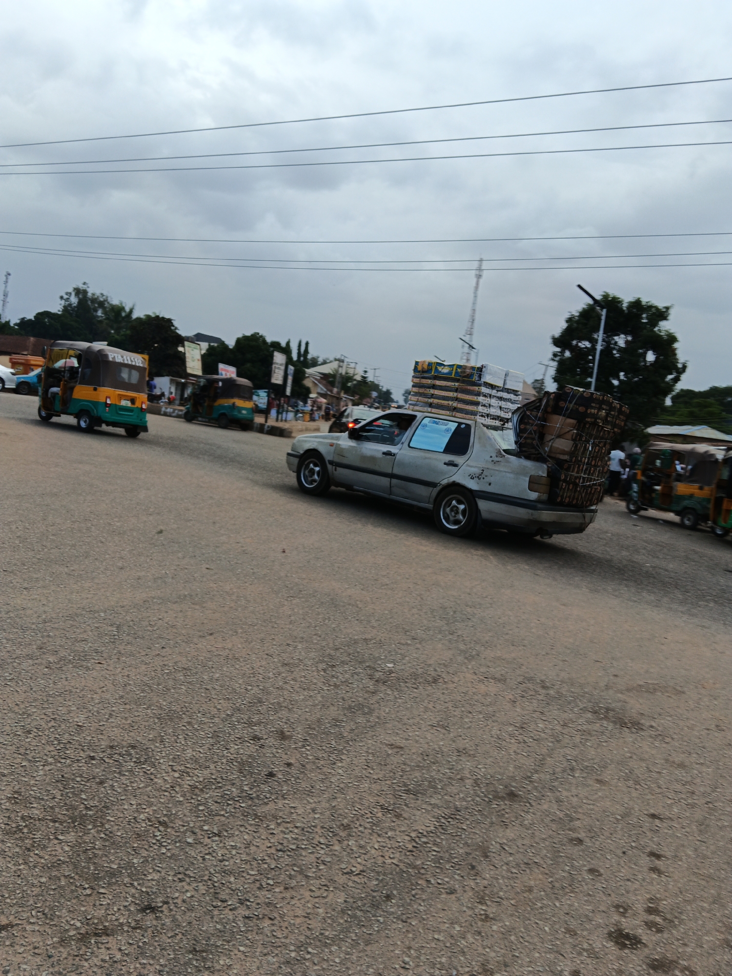 In Bukuru, Nigeria, a silver sedan overloaded with goods travels down a paved road. The car's trunk and roof are laden with boxes and bundles tied with netting. In the background, three-wheeled auto-rickshaws, known as "Keke Marwa," are visible, along with trees, utility poles, and buildings. The sky is overcast, suggesting a cloudy day. The road appears to be a busy thoroughfare in the city.