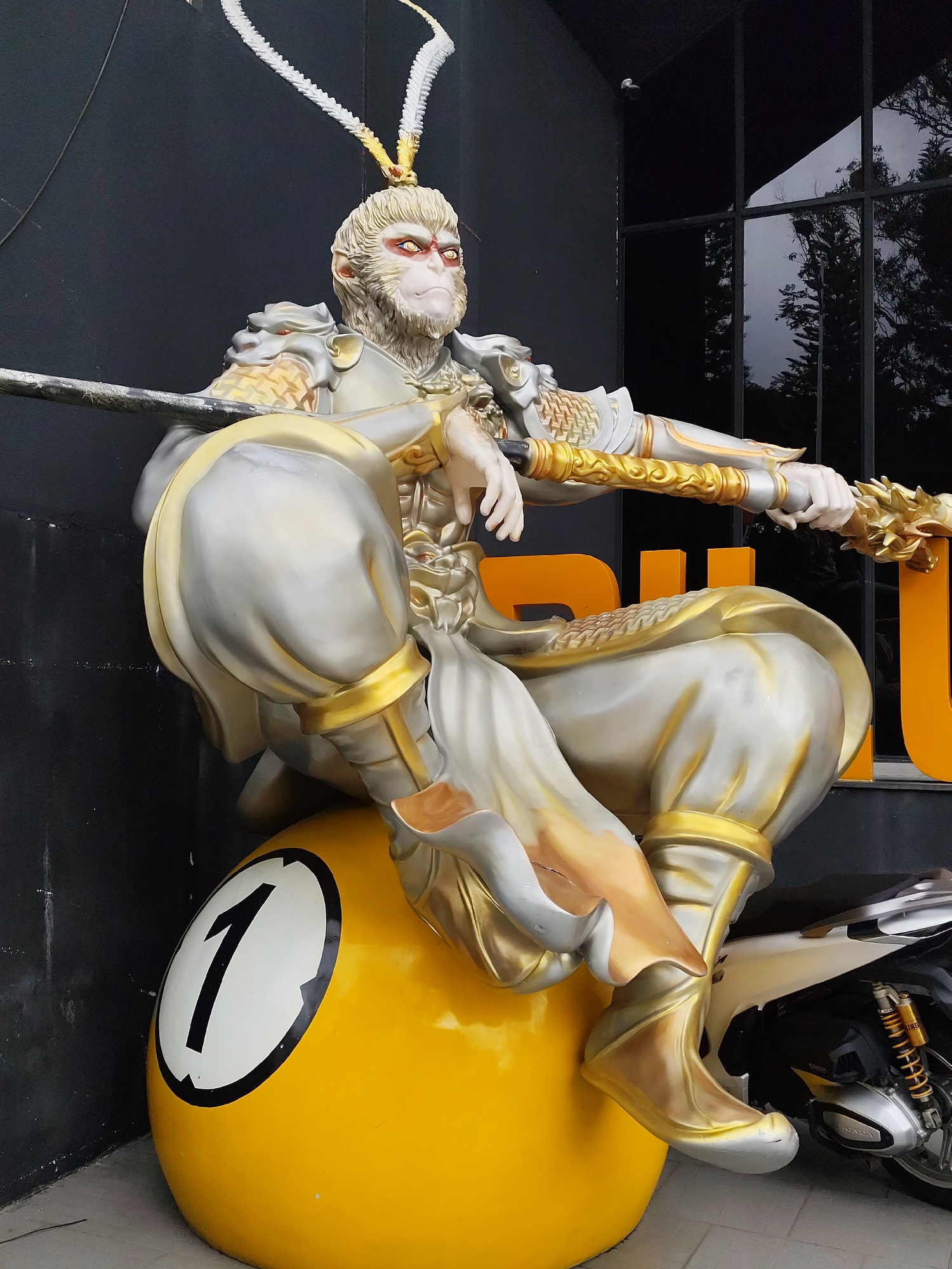 A statue of Sun Wukong, the Monkey King from Chinese mythology, sits atop a yellow ball marked with a black "1" on a white background, located in Da Lat, Vietnam. The statue is silver and gold, dressed in traditional garb and holding a rod. A portion of a motorcycle is visible on the right side of the statue. The statue is positioned in front of a dark, almost black background consisting of a wall and a window.