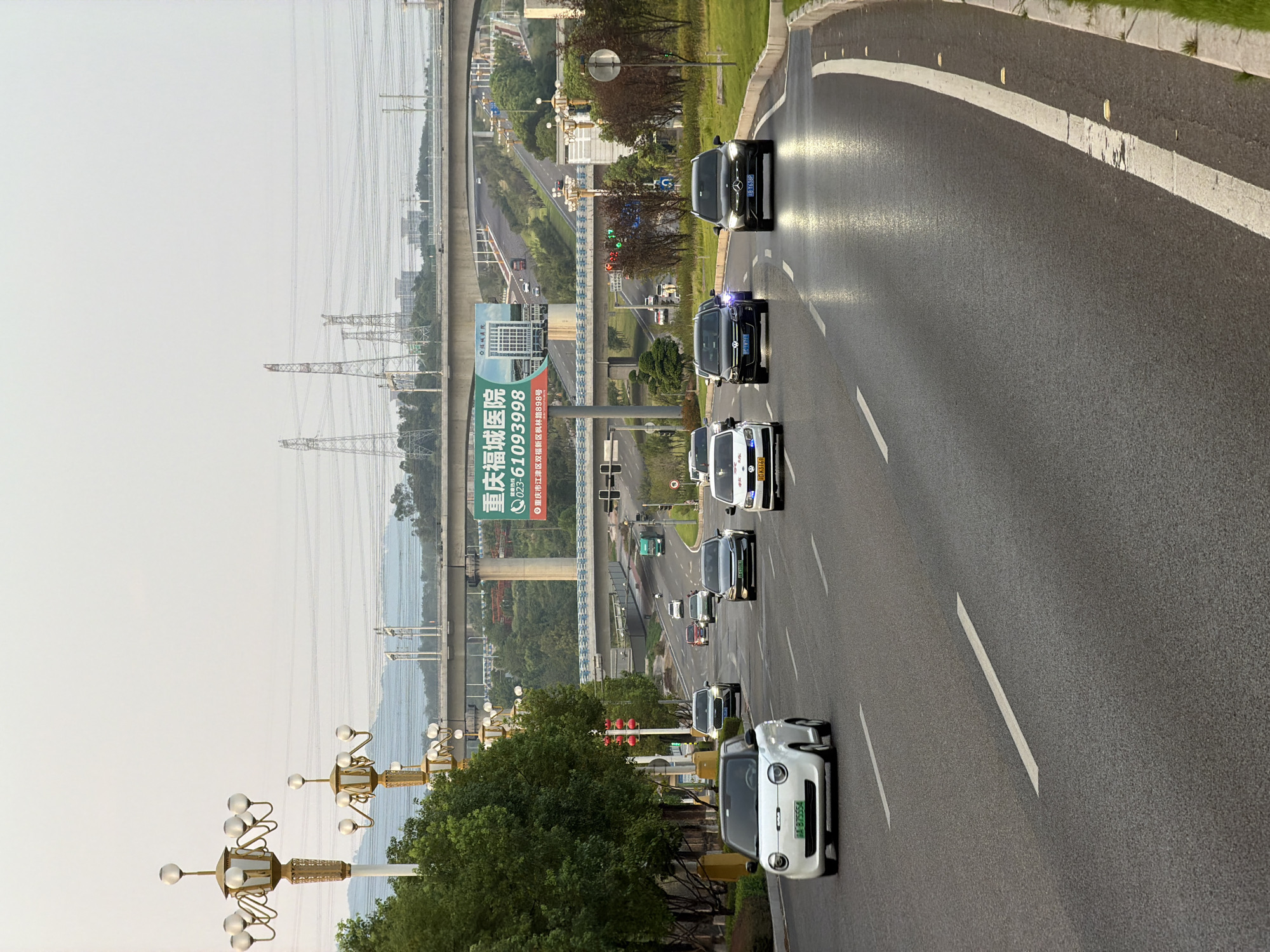 The image depicts a multi-lane road in Jiangjin, China, likely an urban or suburban area given the presence of streetlights, overpasses, and billboards. The time of day appears to be late afternoon or early evening, with soft lighting.
In the foreground, several cars are driving on the road. A small white electric car is in the left lane, followed by a dark sedan. In the right lane, a dark sedan with its headlights on is approaching, and a white sedan is further ahead. Other vehicles are visible in the distance, including a green truck.
The road is well-maintained, with clear lane markings. The surrounding environment includes lush green trees on the left side of the road and more sparse vegetation on the right. In the background, elevated highway structures and power lines are prominent. A large billboard advertising "重庆福城医院" (Chongqing Fucheng Hospital) with a phone number is visible on the right side of the road. The sky is overcast and a light gray.
The scene is a typical view of urban traffic in China, characterized by modern infrastructure and a mix of vehicle types. There are no visible pedestrians or any specific interactions between the vehicles, suggesting a routine flow of traffic.