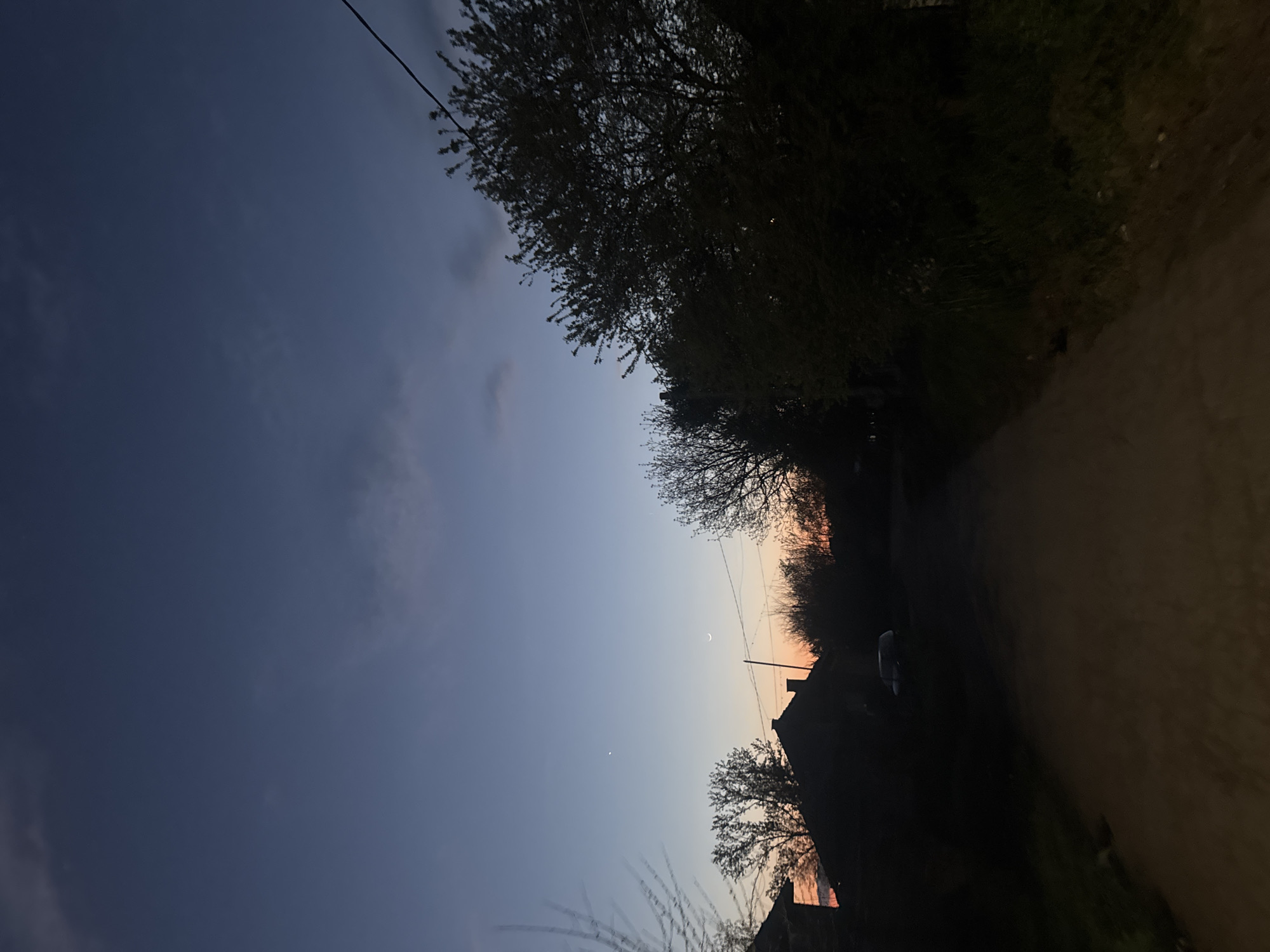 The image shows a rural scene at dusk in Lehchevo, Bulgaria. A narrow dirt road is flanked by dense vegetation on one side and open sky on the other. The sky exhibits a gradient of colors, from a deep blue at the top to a faint orange glow on the horizon. A crescent moon and a small star are visible in the sky. Silhouettes of trees and buildings are visible against the colorful sky. A power line stretches across the top of the frame. The overall lighting is dim, suggesting the photograph was taken during twilight.