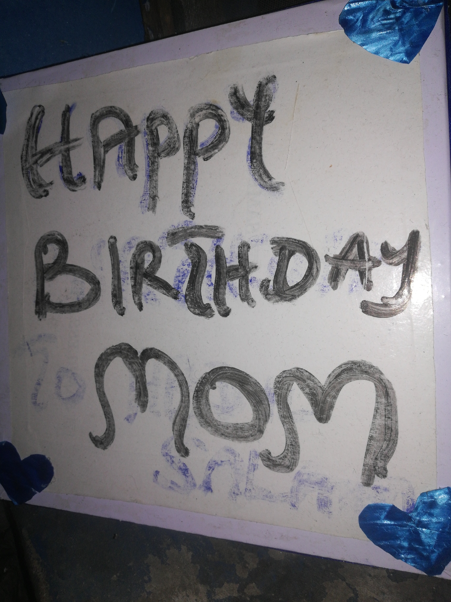A rectangular white board or poster displays the message "HAPPY BIRTHDAY MOM" in large, hand-drawn, thick-stroked capital letters using dark gray or black pigment. Faint blue pen or marker traces are visible beneath the dark lettering. Four blue heart-shaped embellishments are positioned in the corners of the white surface; the upper right heart is made of crinkled metallic blue material, and the lower left heart is solid dark blue. A light lavender border frames the white board. The board rests on a dark, textured surface, likely concrete flooring, with a dark blue object or surface partially visible in the upper background. The scene is observed in Lagos, Nigeria.