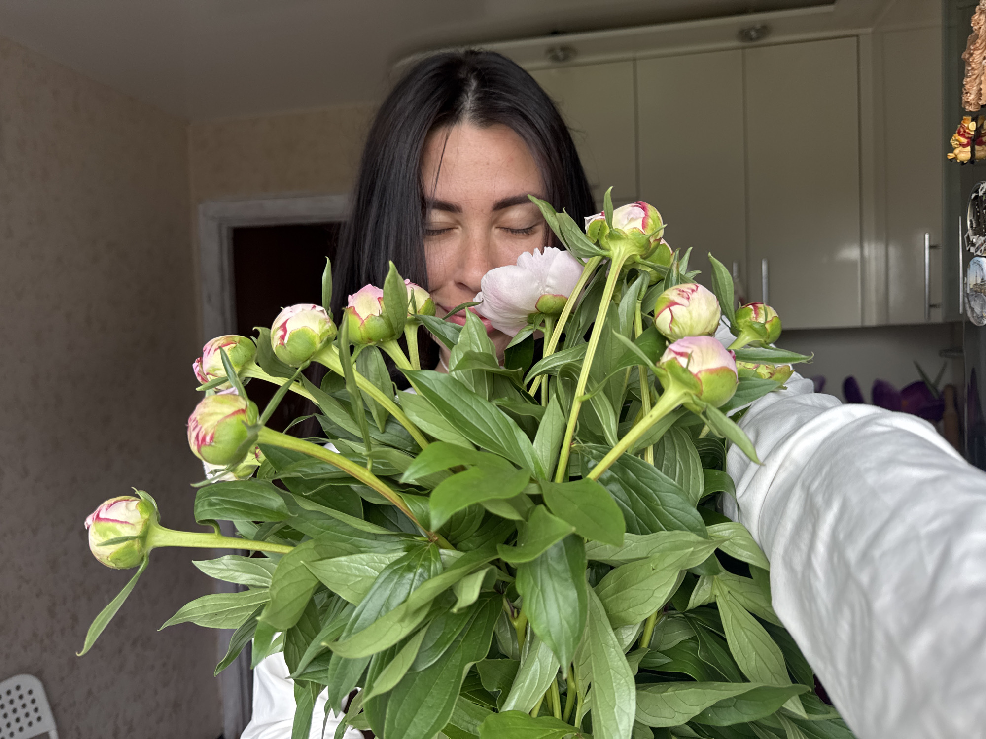 In a domestic interior setting, a woman with dark hair is holding a bouquet of peonies, some of which are in bud and some are blooming with pale pink petals. She is looking down at the flowers with her eyes closed, appearing to smell them. She is wearing a white garment, possibly a shirt or sweater. The background shows a kitchen area with light-colored cabinets and a doorway. A chair is visible in the lower left corner, and some decorative items are attached to a surface on the right. The image appears to have been taken in Chaikovsky, Russia, based on the context.