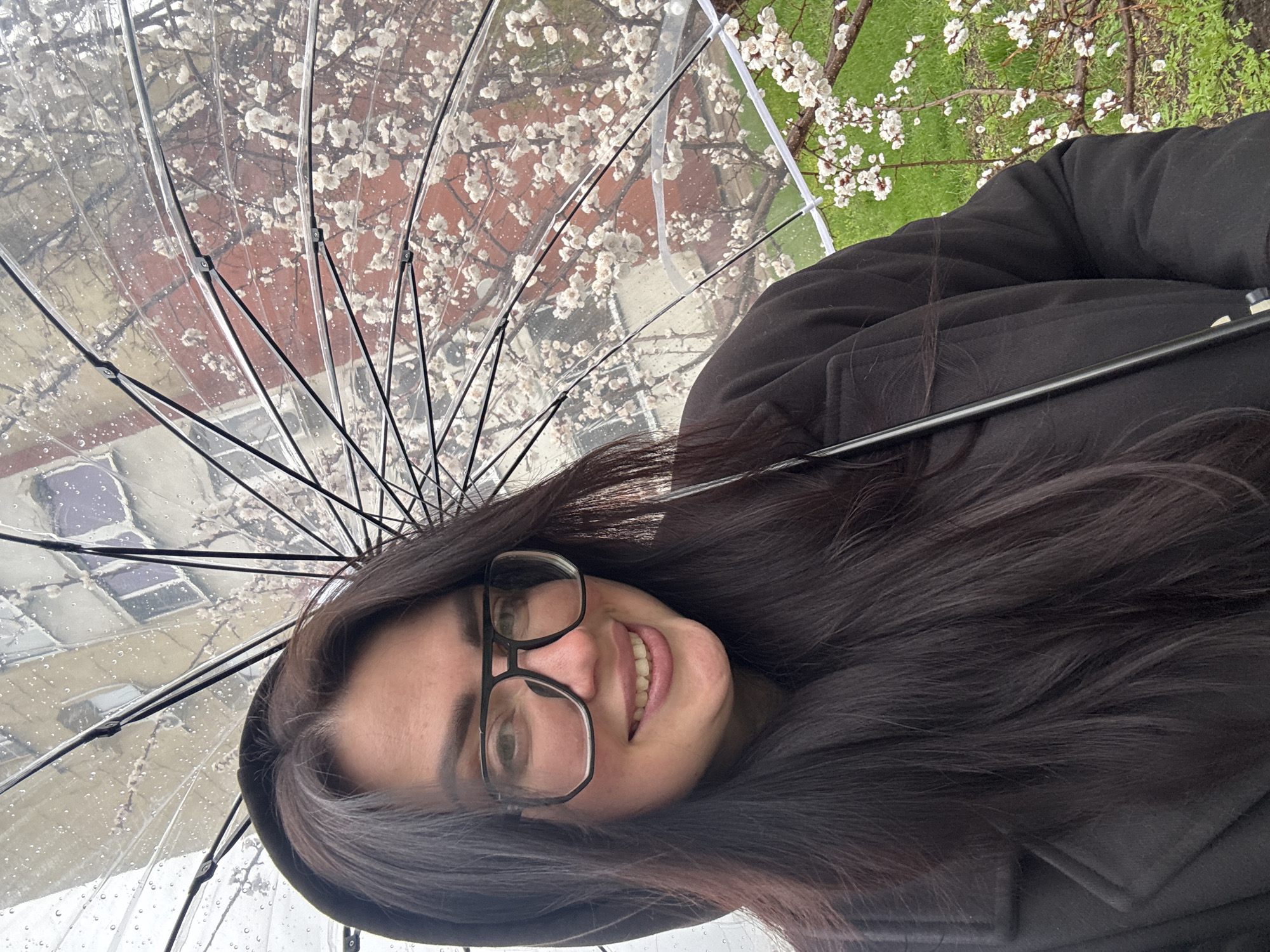 A woman wearing glasses and a dark coat is holding a clear umbrella while standing in an urban environment, likely on a sidewalk. The umbrella shields her from the rain, visible as droplets on its surface. Behind the umbrella, trees with white blossoms suggest it is springtime. In the background, there are buildings. The location seems to be Unknown, Unknown.