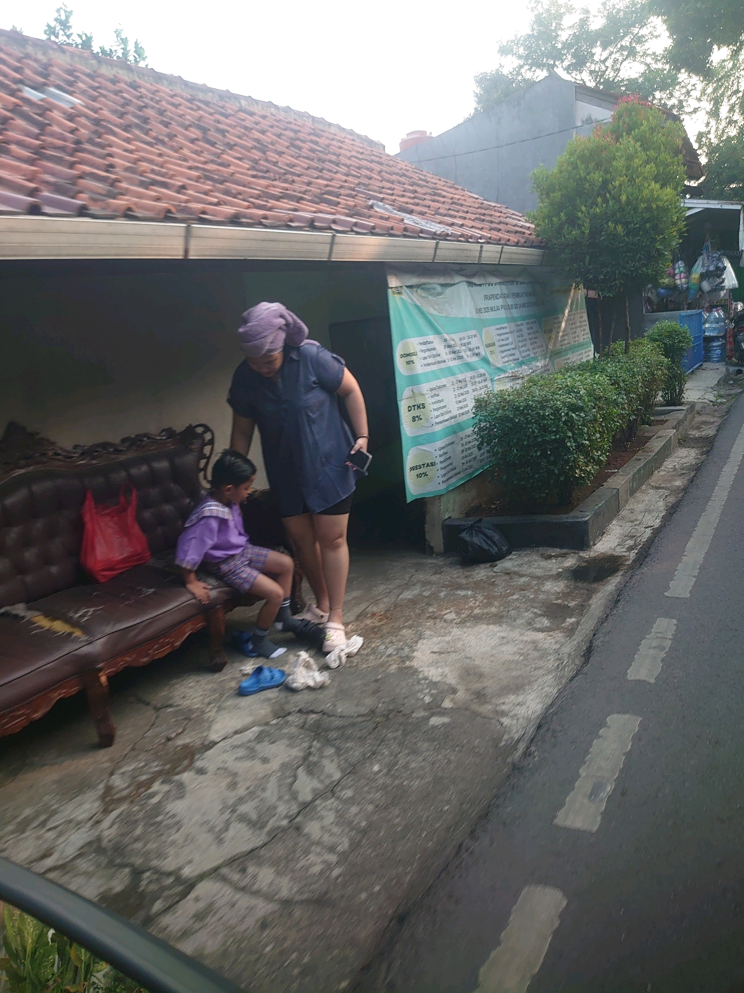 A woman in a blue shirt and purple headscarf stands over a young boy sitting on a brown sofa. The boy is wearing a purple shirt and plaid shorts. A red plastic bag rests on the sofa next to him. The scene appears to be in a residential area, with a tiled roof building and lush green bushes lining a paved walkway. The walkway leads to a street with white dashed lines, suggesting it's a public road. The overall impression is one of a casual, everyday moment taking place outdoors.
