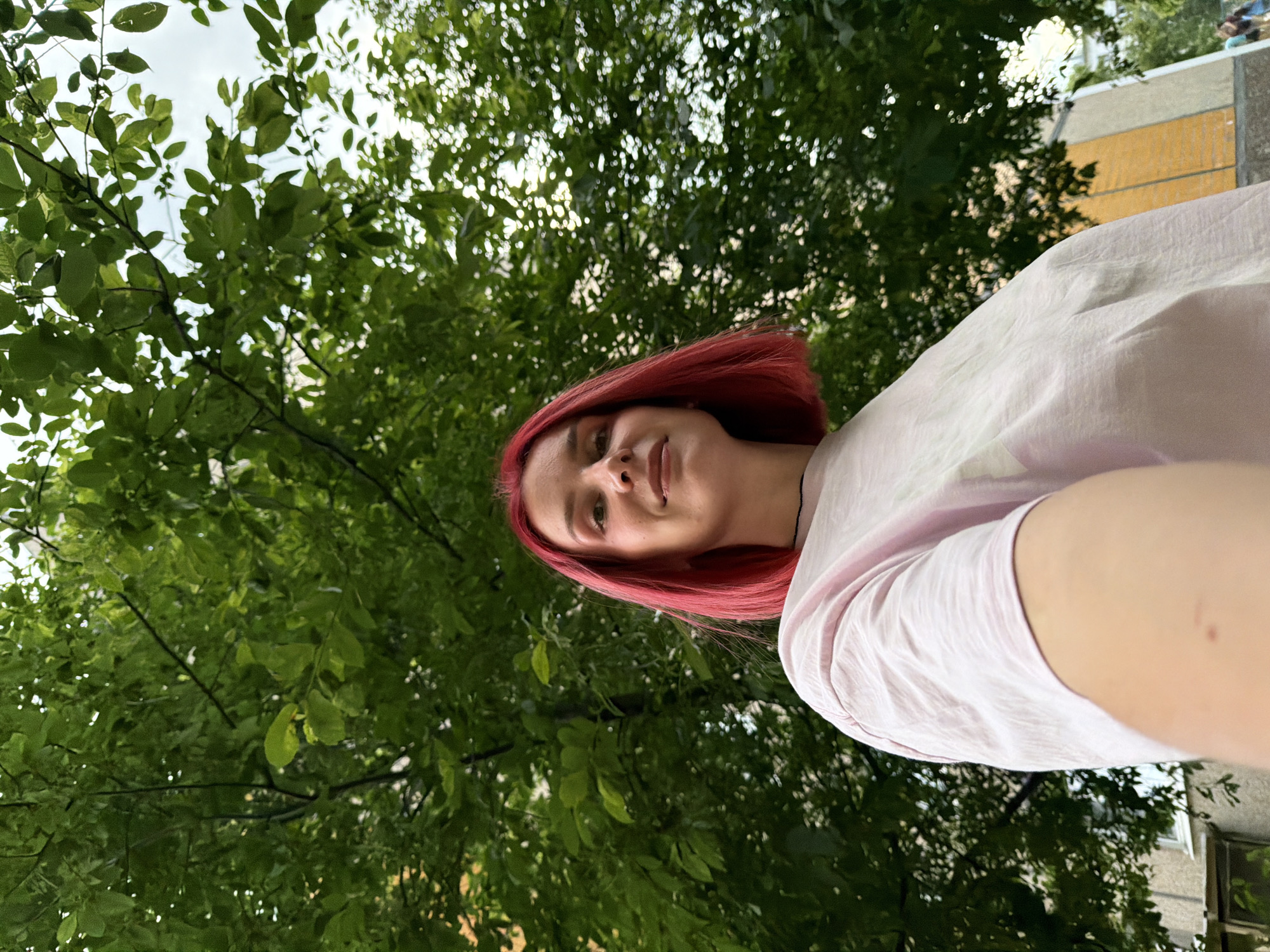 An outdoor, low-angle, medium shot captures a young woman with short, bright red hair. She is wearing a light pink t-shirt. Her face is turned slightly towards the camera, and she has a subtle smile. Behind her, lush green leaves of a tree fill the upper portion of the frame, with glimpses of a building and the sky visible through the foliage. The scene is likely in the city of Chaikovsky, Russia, given the context.