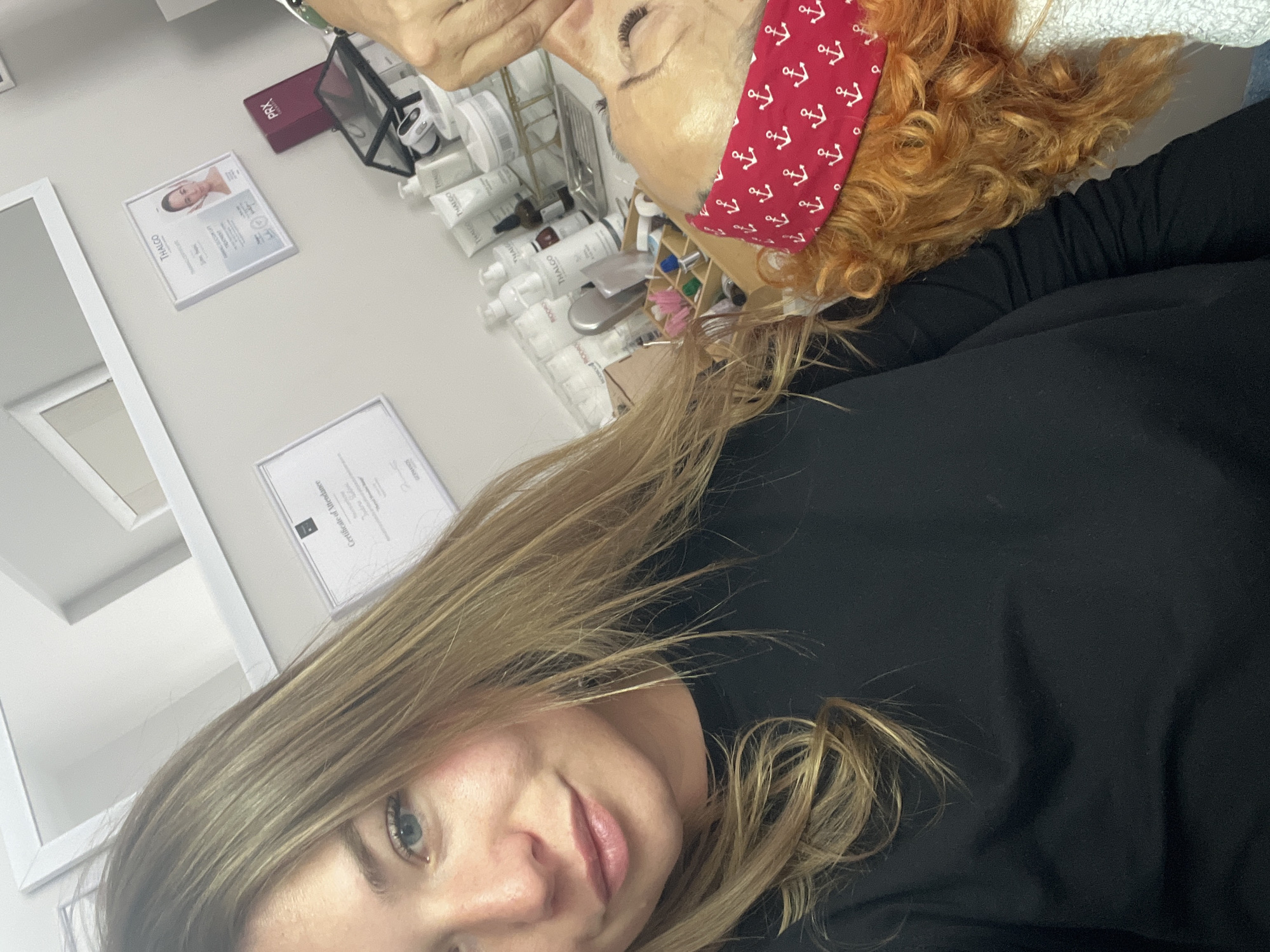 The image, oriented on its side, depicts a selfie taken by a woman with long, light brown hair and blue eyes, wearing a dark top. She looks directly at the camera with a calm, slightly smiling expression. In the background, another person with curly red hair and a red headband patterned with white anchors is visible, lying down and receiving a beauty treatment. Their eyes are closed, suggesting relaxation.
The setting is a professional beauty salon or spa. A counter or shelf is laden with numerous skincare products, predominantly white tubes and bottles. Visible brand names include "THALGO" on many items and "MORIA" on others. A red box with "PRX" is also present, alongside small dark bottles, a silver tool, and a wooden holder containing various brushes and applicators.
Several framed certificates adorn the light-colored wall. One clearly displays "THALGO," a woman's face, and the text "Certificate of Attendance." Another partially visible certificate also includes "Certificate of Attendance" and "GERMANOS." The environment appears clean and professional, consistent with a beauty treatment facility in Budva, Montenegro. No specific time of day or weather is indicated.