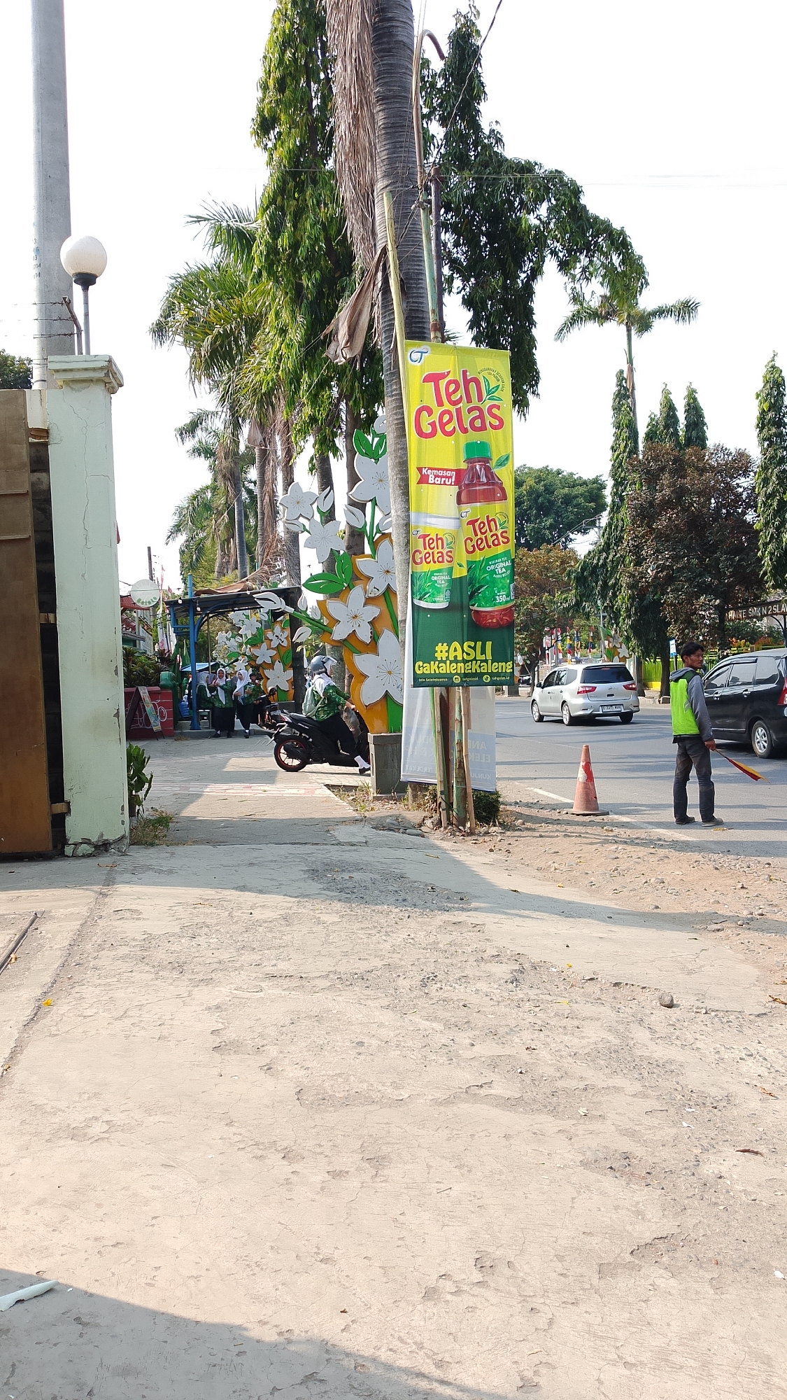 The image depicts a street scene in Procot, Indonesia, on a sunny day. A tall, yellow banner advertising "Teh Gelas" (Glass Tea) is prominently displayed, featuring images of tea bottles and cans. The banner is attached to a pole, and to its left, a white pole with a light fixture is visible.

In the foreground, a paved walkway runs alongside the street. To the left of the walkway, an entrance to a building or property is partially visible, with a weathered, cream-colored gate. Further down the walkway, several individuals are present, some wearing headscarves. A motorcycle is parked on the walkway, with a person riding it.

The street itself is visible in the mid-ground and background. A silver minivan and a black car are driving on the street. A person wearing a bright green vest and dark pants stands on the side of the road, holding a red and yellow object. An orange traffic cone is placed on the street. Tall trees with lush green foliage line the street, some appearing to be palm trees. The sky is bright and clear, indicating good weather conditions. The overall environment suggests a typical urban street in Indonesia, with commercial activity and pedestrian and vehicular traffic.
