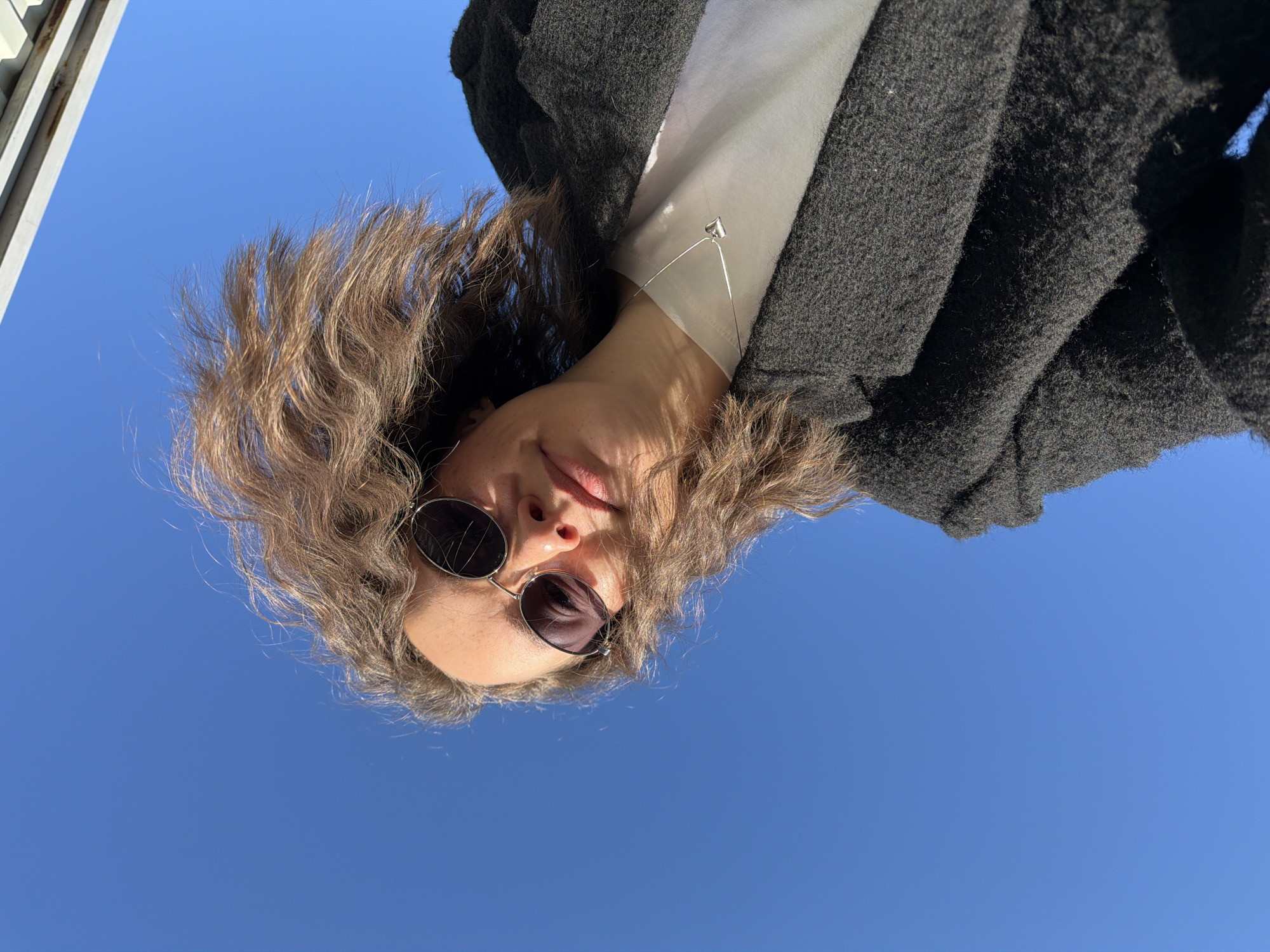 The image shows a person from a low angle looking upwards towards the camera. The person is wearing sunglasses and a dark grey coat over a white shirt or blouse. A silver necklace with a small pendant is visible. The background is a clear blue sky. A partial view of a structure with white vertical and horizontal elements is visible in the upper left corner of the image. There is no way to know if this image was taken in the city of Chaikovsky, Russia.