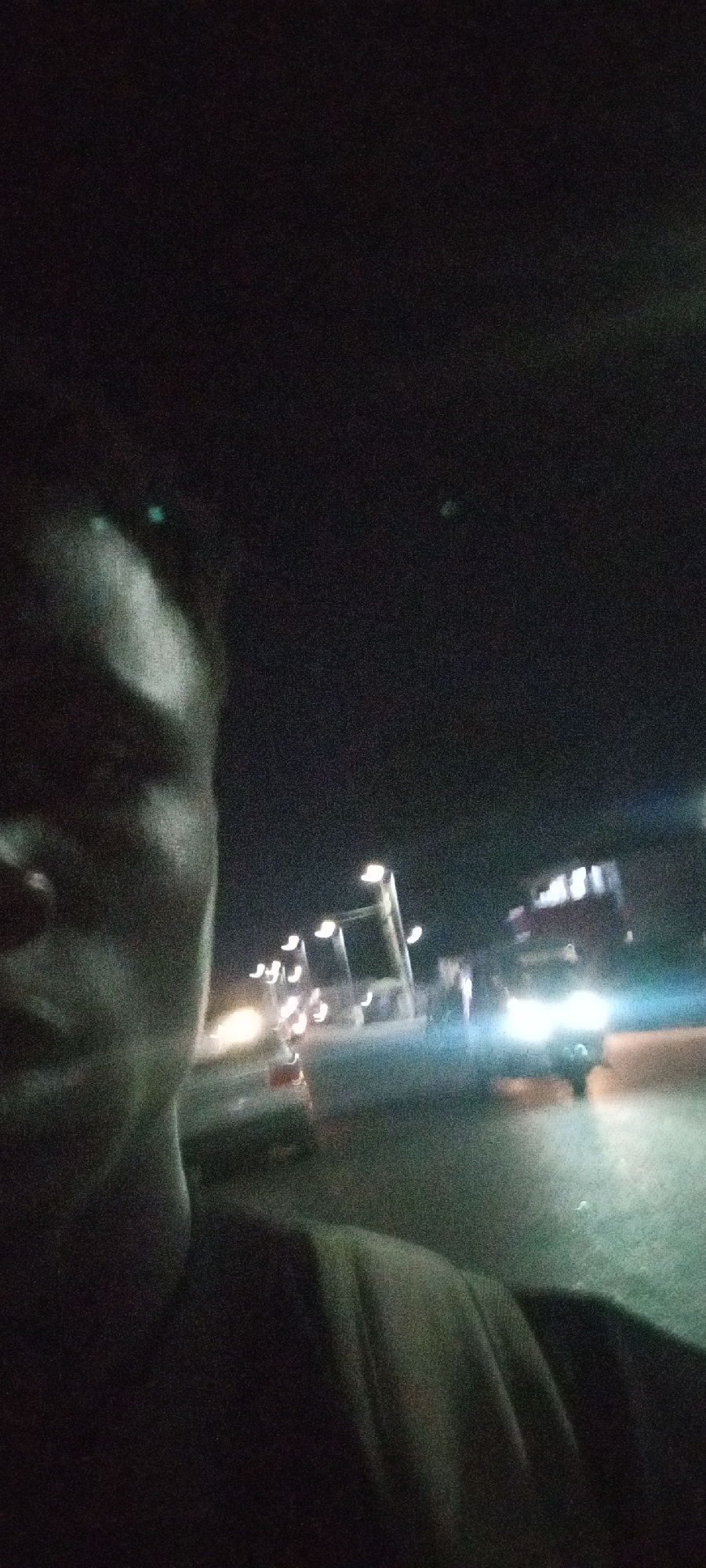 At night, an individual is partially visible on the left side of the frame. Streetlights line the road, illuminating cars and a larger vehicle, possibly a truck, in the background. The location appears to be a road in Jos, Nigeria.