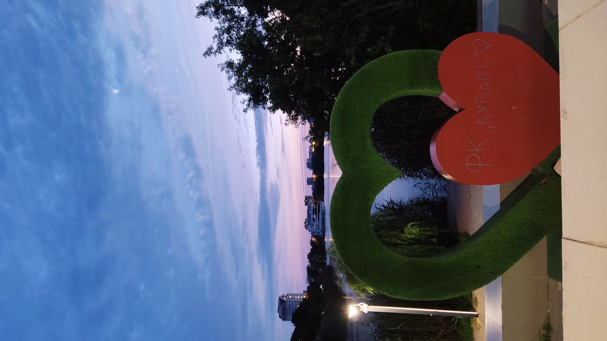 The image shows a waterfront scene at dusk in Krasnodar, Russia. In the foreground, a large green heart-shaped sculpture covered in artificial grass is partially visible. Nestled within the green heart is a smaller red heart sculpture with white Cyrillic text, which reads "ФК, КУБАНЬ" and a small heart symbol, suggesting it relates to the Kuban football club.
Beyond the sculptures, a body of water, likely a river or lake, is visible. On the opposite bank, a city skyline with buildings and trees can be seen. The sky is a mix of deep blue and soft purple hues, indicative of twilight, with some wispy clouds and a sliver of the moon visible. A modern lamppost with a bright light stands to the left of the green sculpture. The overall setting appears to be a park or promenade area along the waterfront.