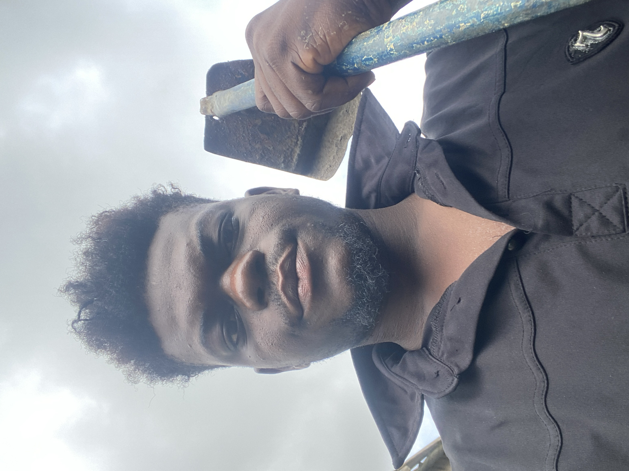 A medium shot depicts a man holding a garden hoe with a blue handle. He faces the camera directly with a serious expression. He has dark skin, curly hair, and a short beard. The man is wearing a black collared shirt. The background consists of a cloudy sky. The location is not discernible in the image.