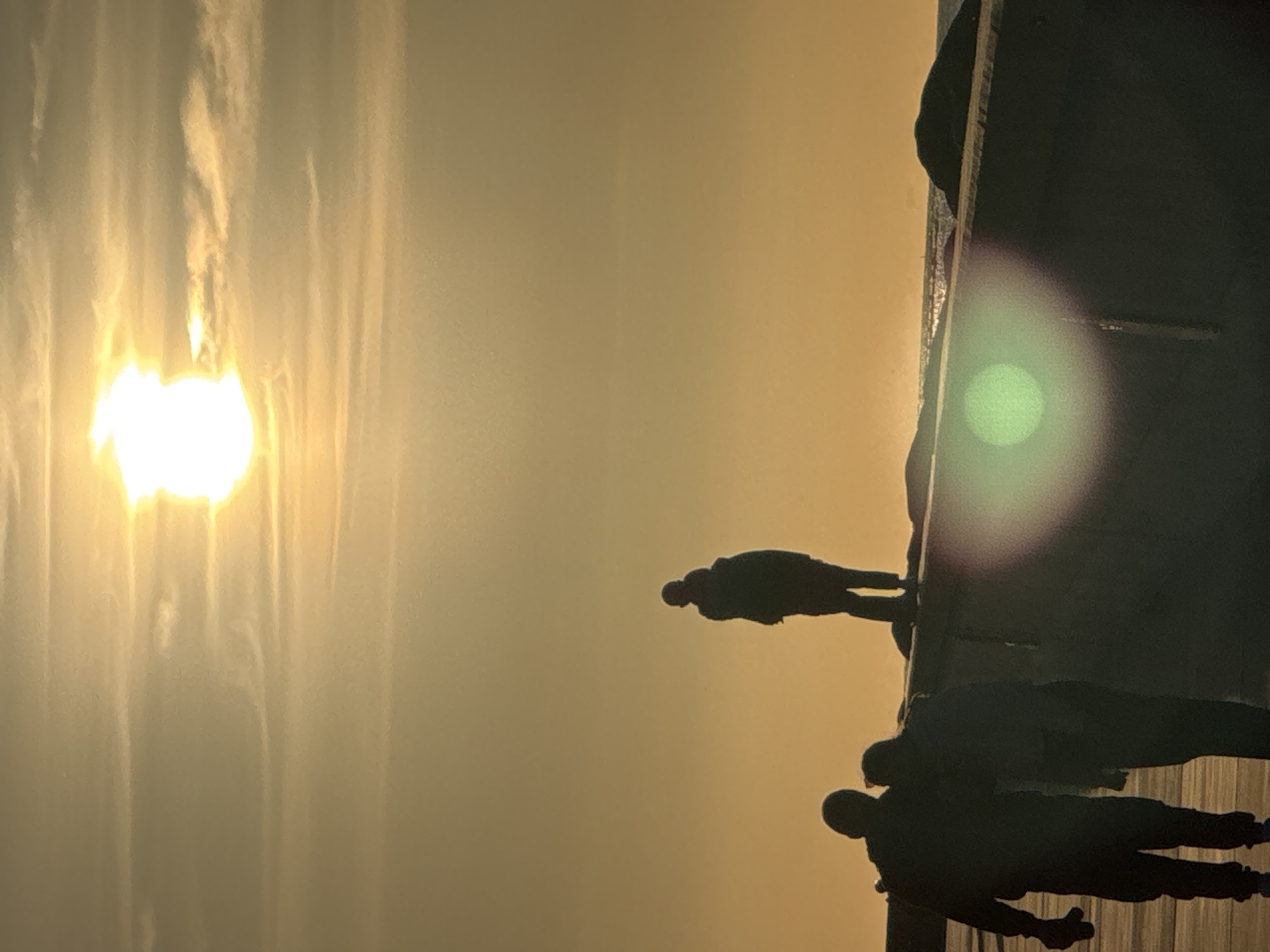 The image shows a sunset over the Baltic Sea, likely taken from the Tallinn coastline in Estonia. Silhouetted against the bright, hazy sun are three figures walking along a seawall. Two figures are walking closely together in the foreground, with one with an arm around the other's shoulders. A third figure stands further away, looking out towards the sea. The sky is a warm gradient of orange and yellow, with wispy clouds. The seawall, made of dark, rough material, curves into the distance, with large rocks visible on its surface. The sea itself is calm and reflective. A green lens flare is present in the foreground, obscuring a small portion of the seawall and the figures.