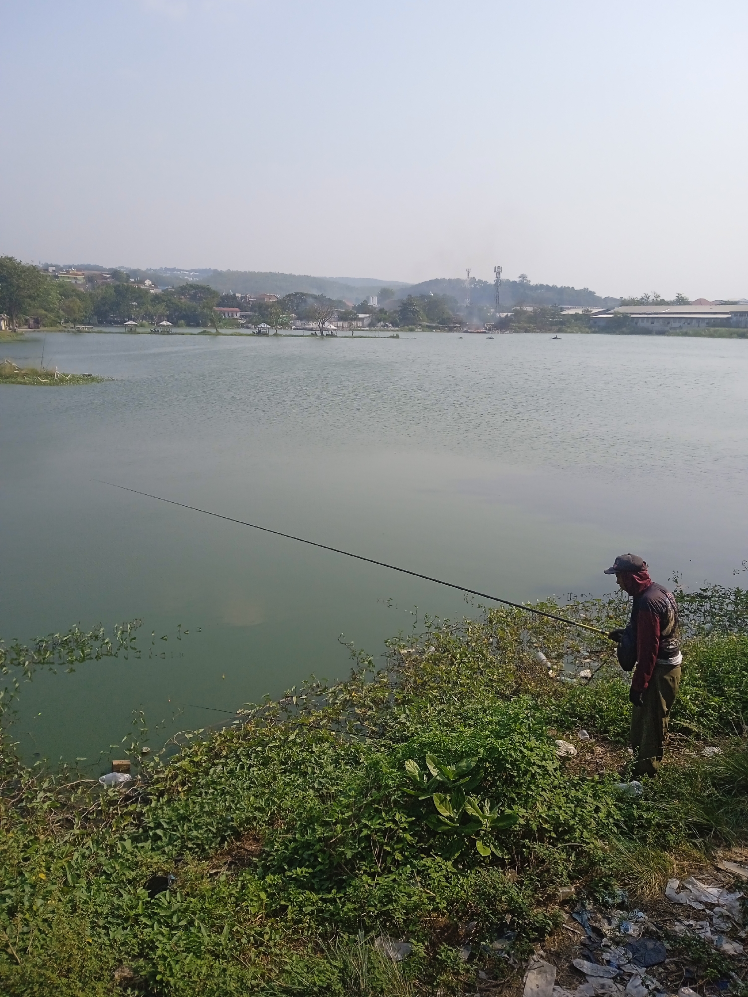 A person is fishing on the shore of a calm, green body of water, likely a lake or reservoir, near Semarang, Indonesia. The person is wearing a dark cap, a red and dark patterned shirt, and green pants, and is holding a fishing rod that extends far into the water. The shoreline is densely covered in green vegetation and appears to have some scattered debris, including plastic bags. In the background, across the water, there is a populated area with houses, buildings, and vegetation on a gentle hill. Several communication towers are visible on the horizon. The sky is clear and light.