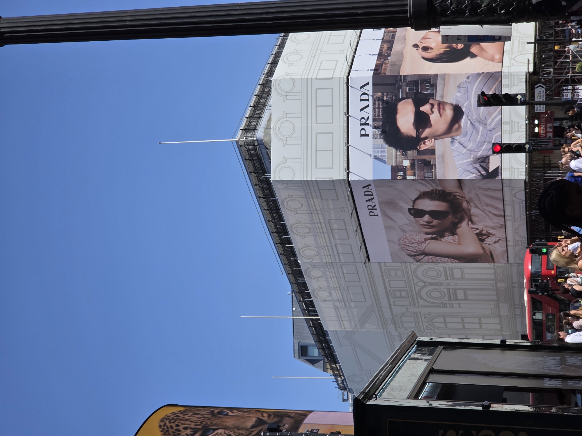 The image shows a view of Piccadilly Circus in London, United Kingdom. The scene features the iconic advertising screens, prominently displaying Prada advertisements. A red traffic light is visible, indicating stopped traffic. Pedestrians are present in the background. Clear blue sky.