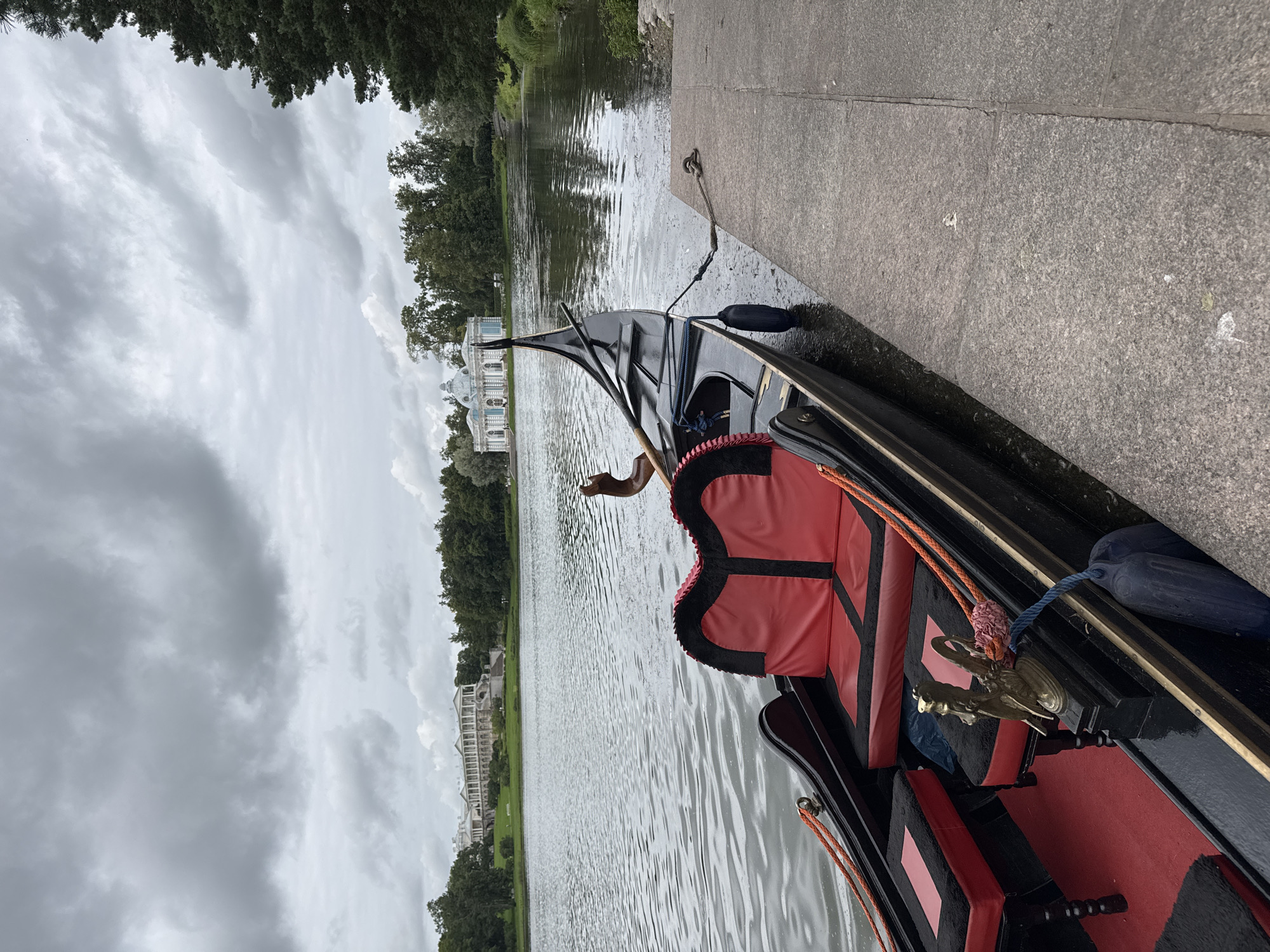 A gondola is docked at a stone quay on a body of water under a cloudy sky. The gondola is black with red and black upholstery and is tied to the quay with a rope. In the background, across the water, is a long, white classical-style building with many columns, possibly a palace or museum. To the right of that building is a smaller, light blue domed structure. Dense green trees line the far shore, and a large evergreen tree is visible on the right side of the image. The water is calm and reflects the overcast sky. The overall scene appears to be a park or estate. The location is likely the Catherine Park in Pushkin, Russia, given the architecture and style of the park.