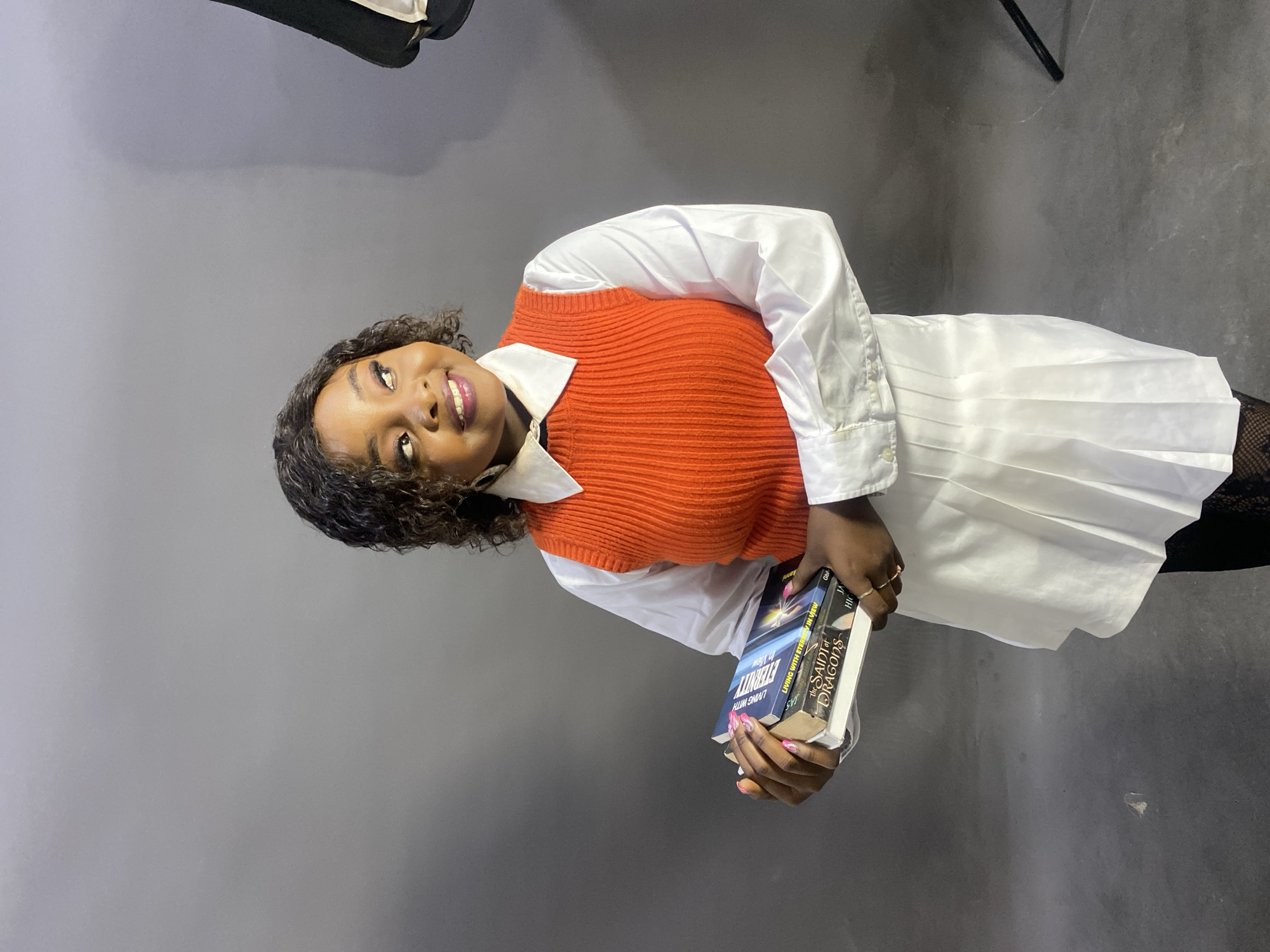 A young woman stands in front of a gray background, likely in a studio setting, holding two books. She is wearing a white button-down shirt with the sleeves rolled up, topped with an orange knitted vest. She has short, curly hair and is looking up and smiling. Her fingernails are painted pink. The books she holds are titled "Living with Eternity in View" and "The Saint of Dragons." She wears black stockings with a fishnet pattern. The scene is likely indoors and in Jos, Nigeria based on the subject's appearance. A lighting apparatus is visible at the top of the frame.