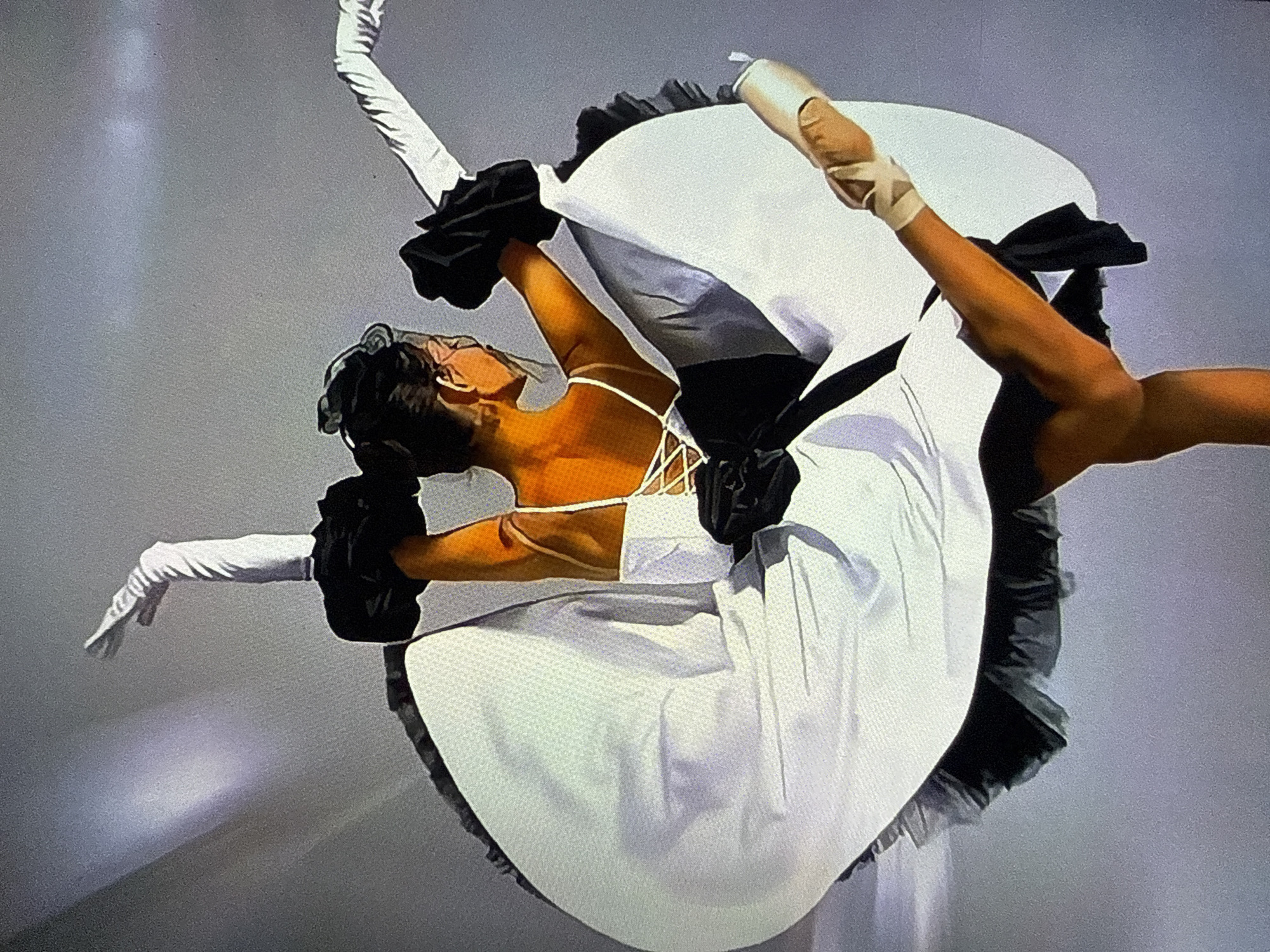 The image depicts a female ballet dancer captured from behind, positioned in a dynamic, mid-turn or arabesque pose. Her body is twisted, with her head turned to look over her right shoulder. Her right arm is raised high, extended upwards, and her left arm is extended outwards to the side. Both hands are covered by long white gloves. Her right leg is extended high and backward, with her foot en pointe in a light-colored ballet shoe secured with ribbons around the ankle. The supporting left leg is largely obscured.
She wears a two-tone costume: a white top with thin spaghetti straps visible over her shoulders and a criss-cross lacing detail on the back. Attached to her upper arms are voluminous black ruffled elements, resembling puffed sleeves. The skirt of her costume is wide and flowing, primarily white, billowing outwards around her lower body, indicative of motion. The underside or trim of the skirt appears black, possibly ruffled or layered. A black sash or band is visible around her waist, and a black ruffled detail is present at the base of the laced bodice. Her hair is dark and styled in a bun at the back of her head, secured by a black accessory. Her skin tone is warm.
The background is a plain, light grey or off-white surface, devoid of any discernible features or markings. A subtle, soft, diagonal beam of light, with a faint purplish hue, is visible in the upper left quadrant, suggesting stage lighting. The specific location, including the city of Varna, Bulgaria, cannot be determined from the visual information present in the image.