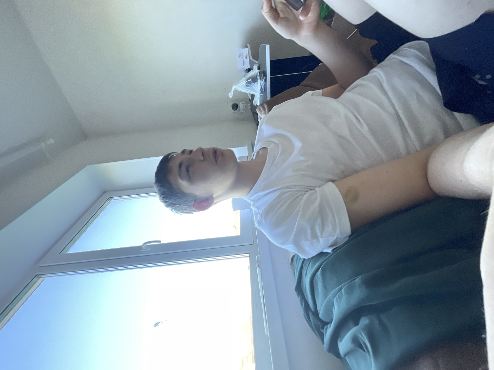A young person, appearing to be male with short, dark hair, is lying down indoors, looking upwards with a neutral expression. They are wearing a white t-shirt and dark green pants or a blanket. There is a bruise visible on their right arm.
The setting appears to be a room with a large window that lets in bright daylight, suggesting it is daytime. The window offers a view of a clear blue sky, indicating good weather. In the background, a white desk or shelf is visible with some items on it, including what looks like a security camera. The room has light-colored walls and ceiling. The overall impression is of a casual, indoor environment. The image is taken from a low angle, looking up towards the subject and the window.
No text is visible in the image. The location context is Красные Баки, Russia.