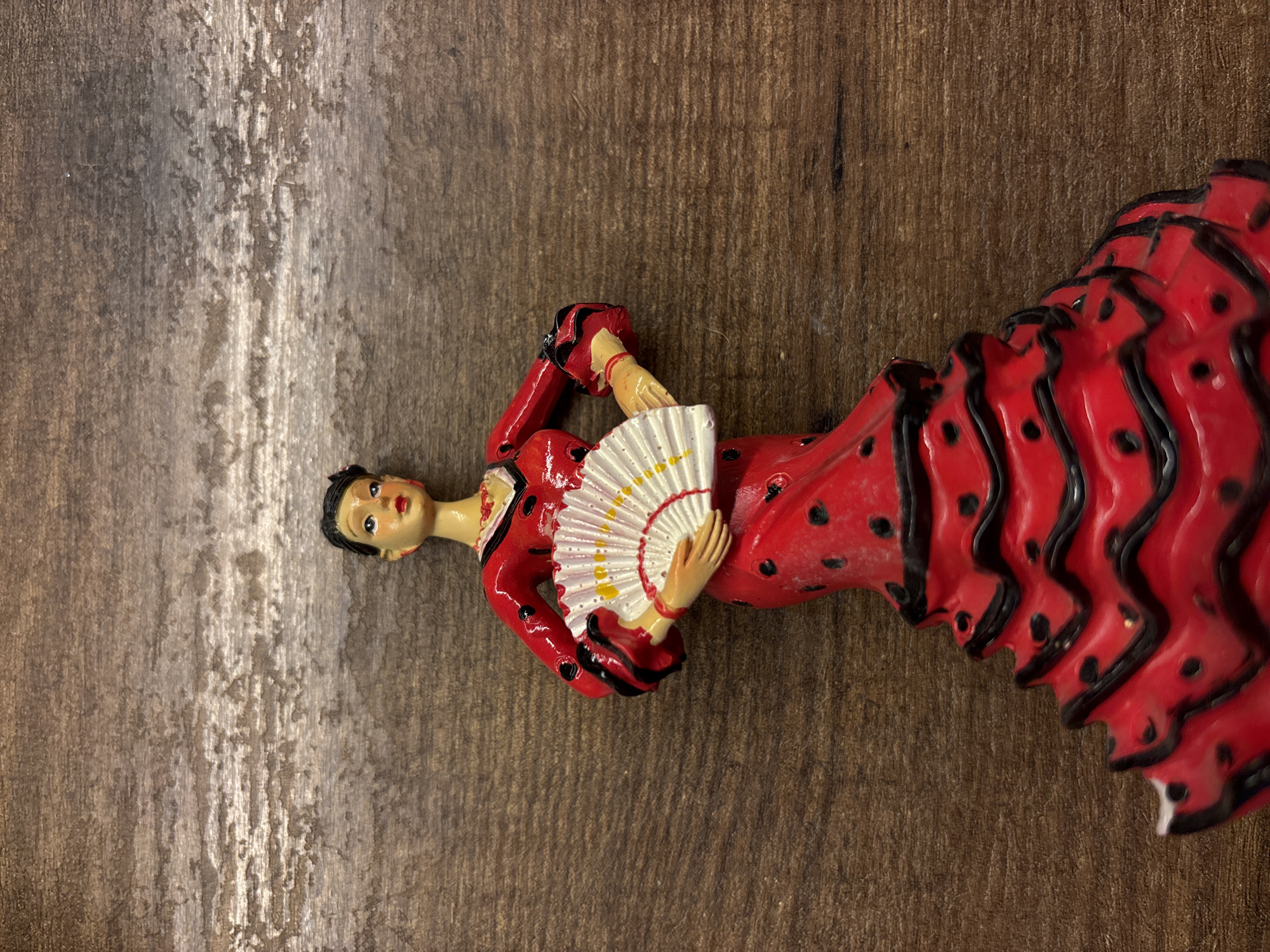The image presents a top-down, close-up view of a decorative female figure, reminiscent of a traditional Spanish flamenco dancer or doll, resting on a textured wooden surface. The figure is prominently dressed in a vibrant red gown featuring multiple black ruffled tiers on the skirt and black polka dots throughout. She wears a red top with black cuffs and trim. Her complexion is fair, her hair is black and styled, and her face is painted with red lips and eyes gazing slightly upwards. The figure holds a white hand fan with subtle yellow markings.
The wooden surface beneath the figure displays a distressed, rustic texture with varying shades of brown and grey, suggesting a worn or aged appearance. The figure is positioned horizontally across the frame. There is no discernible activity taking place, as it is a still life of an object. No text is visible in the image. While the figure's attire evokes Spanish culture, the context provided states the image was captured in Saint Petersburg, Russia.