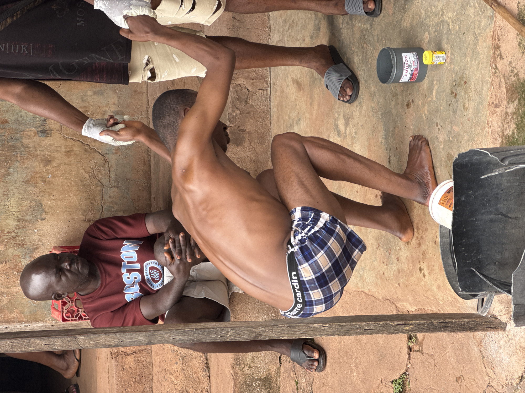 The image shows three men and a bucket. One man, wearing a maroon t-shirt with "BOSTON" written across it, sits on steps. Another man, wearing a black t-shirt and ripped beige pants, stands and holds the arm of a third man. This third man, wearing plaid boxers, crouches down and appears to be reaching for something in the bucket. The bucket is black and appears to contain some white substance. A small yellow bottle and a larger grey container are also on the ground near the bucket. The environment appears to be outdoors, possibly in a courtyard or an alley, with a rough concrete floor and a textured, aged wall in the background. The style of the building and the clothing of the individuals suggest a setting in Enugu, Nigeria.