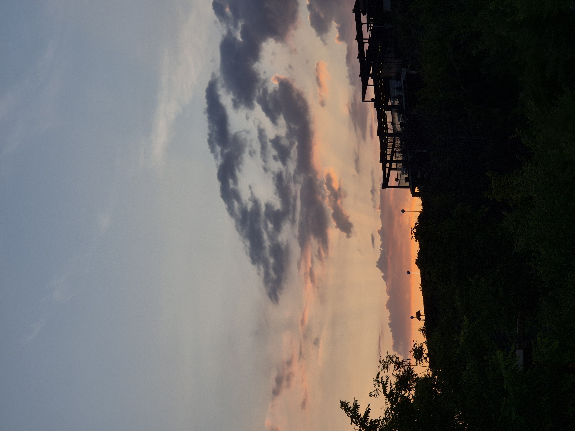 This media file presents a scenic landscape photograph captured during sunset or dusk in Glavatartsi, Bulgaria. The sky dominates the upper two-thirds of the frame, displaying a vivid gradient of colors. On the left, the sky is a soft, pale blue-grey, transitioning towards the right into striking shades of orange, pink, and purple, characteristic of a setting sun. Several clouds are visible; darker, denser grey clouds are scattered across the upper-left and central sky, providing a contrast to the lighter, more ethereal clouds illuminated by the warm sunset hues on the right.
Along the horizon, which forms the lower third of the image, a dark silhouette of the landscape is visible. On the left, a dense cluster of trees and foliage creates an irregular, dark outline. In the central portion of the horizon, a series of low-rise structures, possibly buildings, a resort complex, or attached units, are silhouetted against the vibrant sky. Some faint internal or decorative lights are subtly visible within these dark forms. Several streetlights on tall poles punctuate the horizon line, emerging above the silhouettes, especially towards the right side, indicating a developed area. The immediate foreground, filling the bottom of the image, is a dark, undifferentiated mass of foliage.
The scene captures the tranquil beauty of the transition from day to night. There is no specific activity depicted, and no visible text. The overall impression is one of peaceful natural beauty in a developed or semi-developed setting.