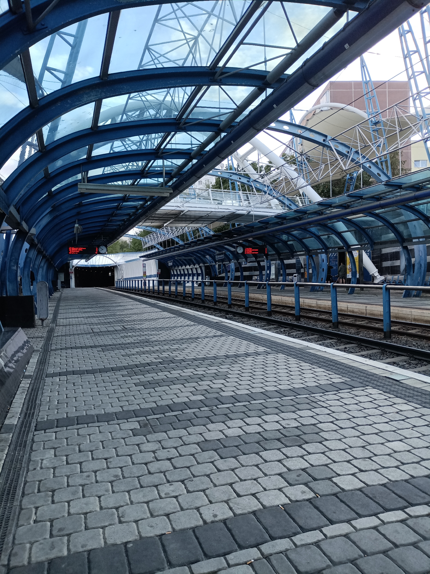 The image depicts an empty tram station platform in Prague, Czechia. The platform is paved with grey and black interlocking stones arranged in a patterned formation. A blue, curved canopy with a glass roof extends over the platform, supported by blue metal beams. Train tracks run alongside the platform. In the background, there is a white structure with a complex metal framework and a large white awning, suggesting further urban development or infrastructure. A digital display board with red text is visible under the canopy, indicating information about train departures. The scene is devoid of any people.