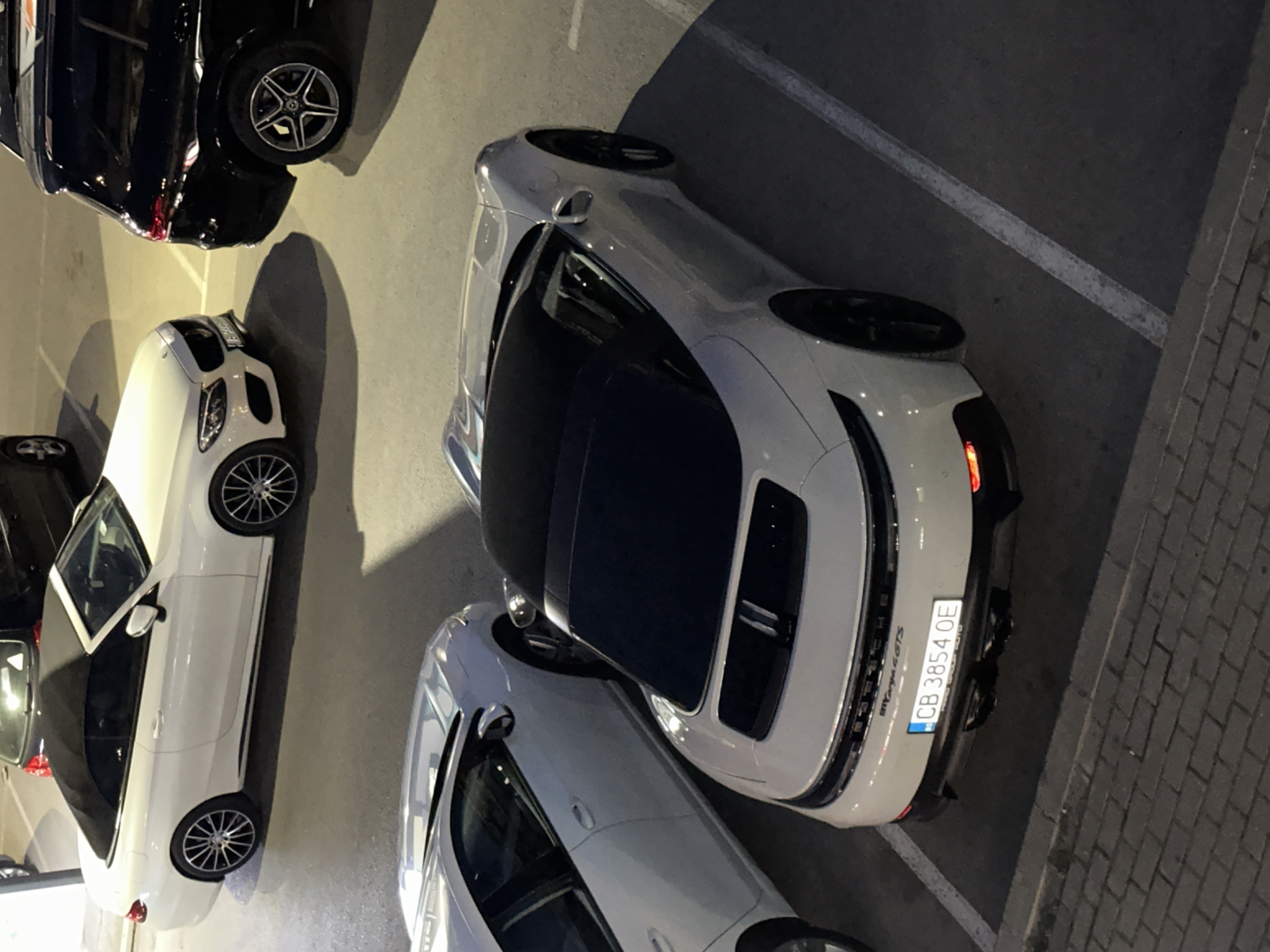 The image shows an overhead shot of a parking lot in Varna, Bulgaria, featuring several parked cars. In the foreground is a white Porsche 911 Targa 4 GTS with license plate "CB 3854 OE" which is registered in Varna. Behind the Porsche is another white convertible Mercedes-Benz, and further back a black Mercedes-Benz SUV is visible. The parking lot is lined with white parking space markers. The surface of the parking lot is asphalt, with brick paving visible at the far right of the frame. The lighting suggests it is nighttime.