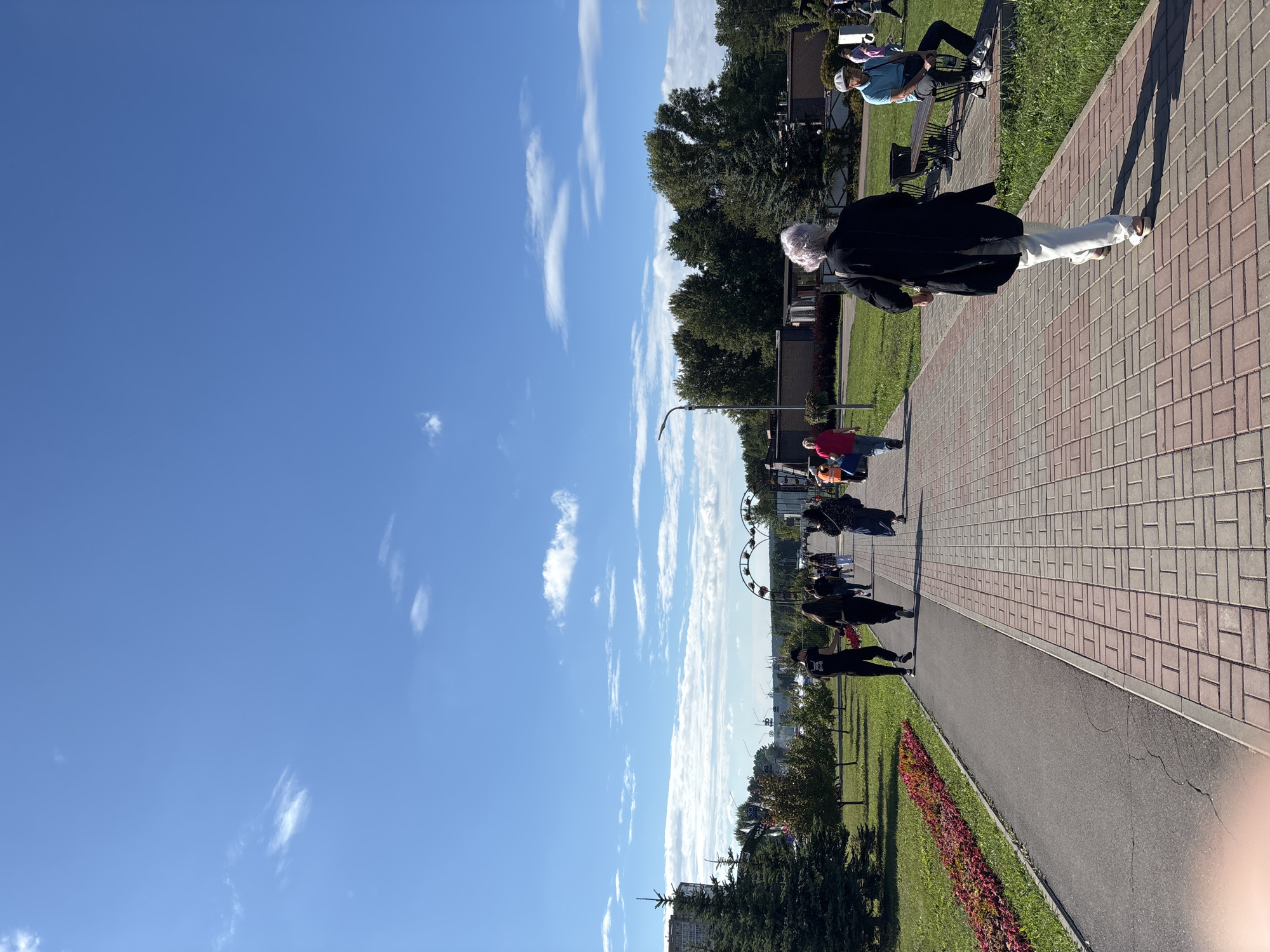 The image captures a sunlit outdoor scene along a paved walkway in Naberezhnye Chelny, Russia. The walkway, constructed with red and grey interlocking bricks, is bordered by a grassy area with flowers and trees. Several people are walking along the path, some are strolling casually, while others appear to be walking with purpose. In the background, there are trees, buildings, and possibly amusement park structures, suggesting a public park or recreational area. The sky is blue with scattered white clouds, indicating fair weather. A woman in a black coat and white pants is walking away from the camera on the paved path. On the right side of the path, a man wearing a blue shirt and a cap is sitting on a bench. The overall scene conveys a peaceful, public gathering on a bright day.