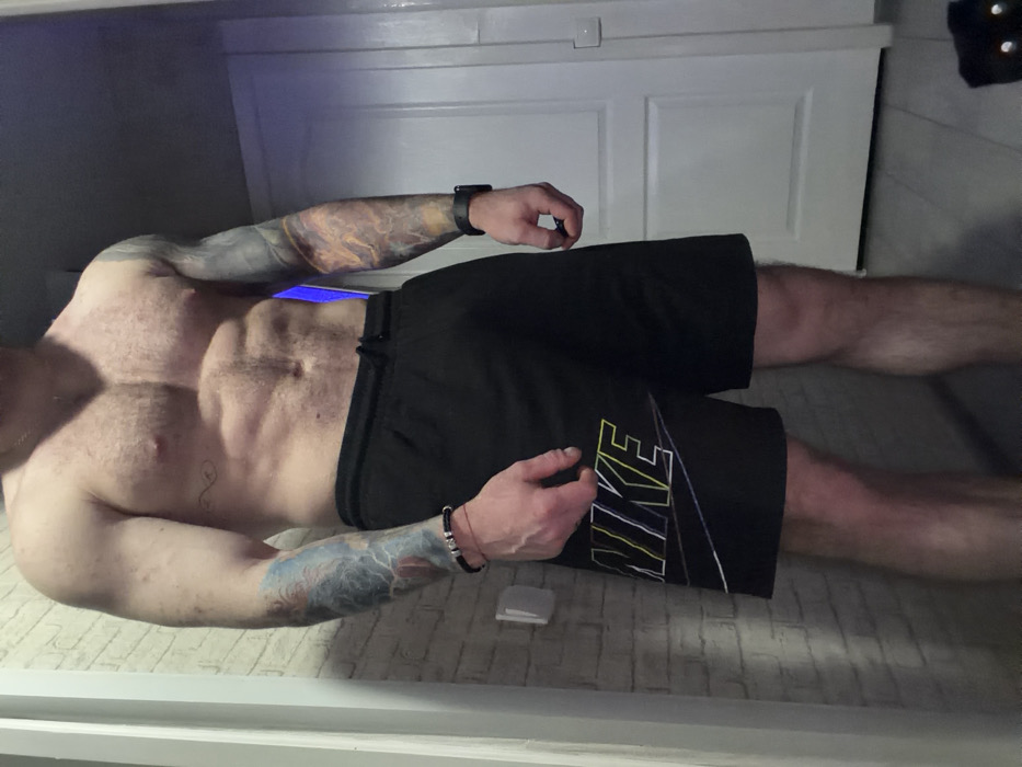 A shirtless man lies inside an open freezer. He wears black Nike shorts. Tattoos cover both arms. He has a watch on his left wrist and a beaded bracelet on his right. The freezer's interior is white with blue light near the top. The man's muscular torso and hairy legs are visible against the textured freezer floor.