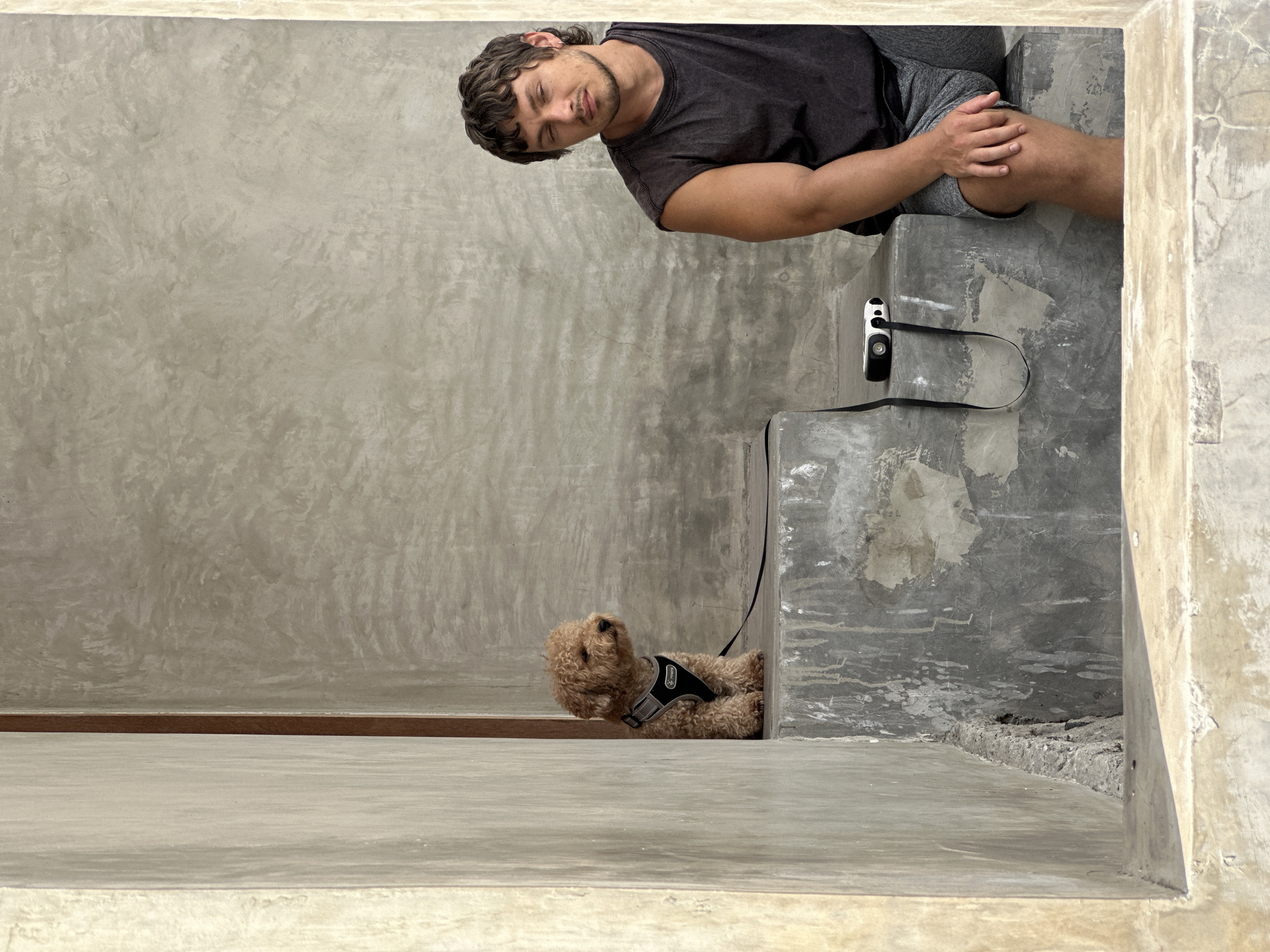 A small, light brown poodle wearing a black harness sits on a concrete ledge to the left of the frame. To the right, a person with dark, curly hair and wearing a sleeveless dark t-shirt and grey shorts sits on a lower concrete ledge, looking down with their eyes closed. A small white camera with a black strap rests on the ledge between the dog and the person. The background is a textured grey concrete wall. The foreground shows a concrete ledge with a rough, aged texture. The overall setting appears to be an outdoor or semi-outdoor urban environment in North Kuta, Indonesia, characterized by raw concrete architecture.