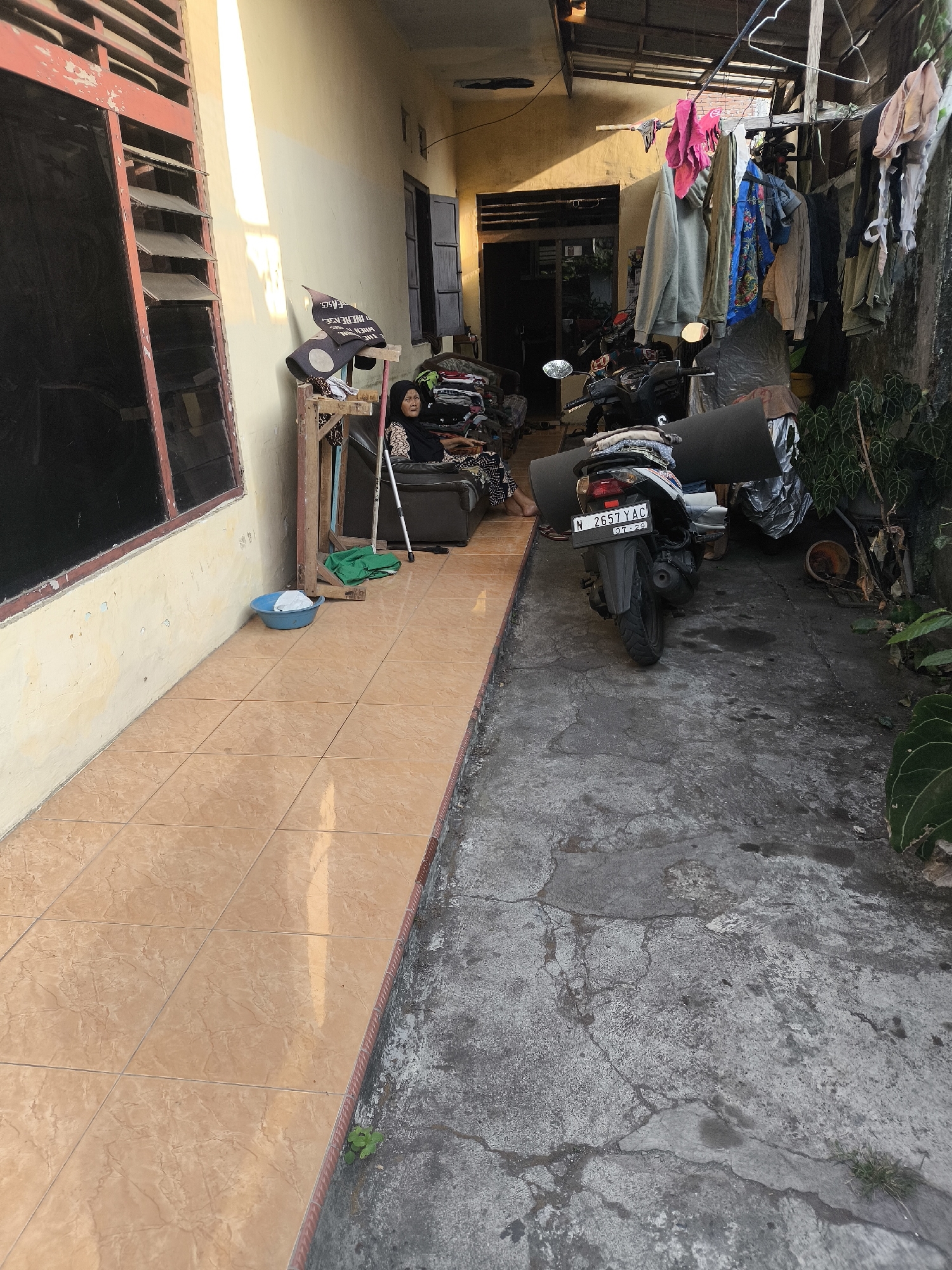 The image depicts an outdoor alleyway in a residential area of Citrodiwangsan, Indonesia. Sunlight illuminates the left side of the scene, casting long shadows. A woman wearing a black hijab and a patterned dress is seated on a dark-colored sofa to the left of the image. A walking stick leans against the sofa. In front of the sofa, a wooden stand holds various items, including what appears to be a black fabric with white lettering. A blue plastic basin is on the tiled walkway in the foreground. A motorcycle is parked in the alleyway. Clothes are hanging on a line overhead, suggesting laundry is being dried. The ground in the alleyway is made of concrete, showing signs of wear and tear, with cracks and some scattered debris. To the right, greenery is visible, including the leaves of a plant. The overall impression is one of a lived-in, somewhat cluttered, and functional outdoor space.