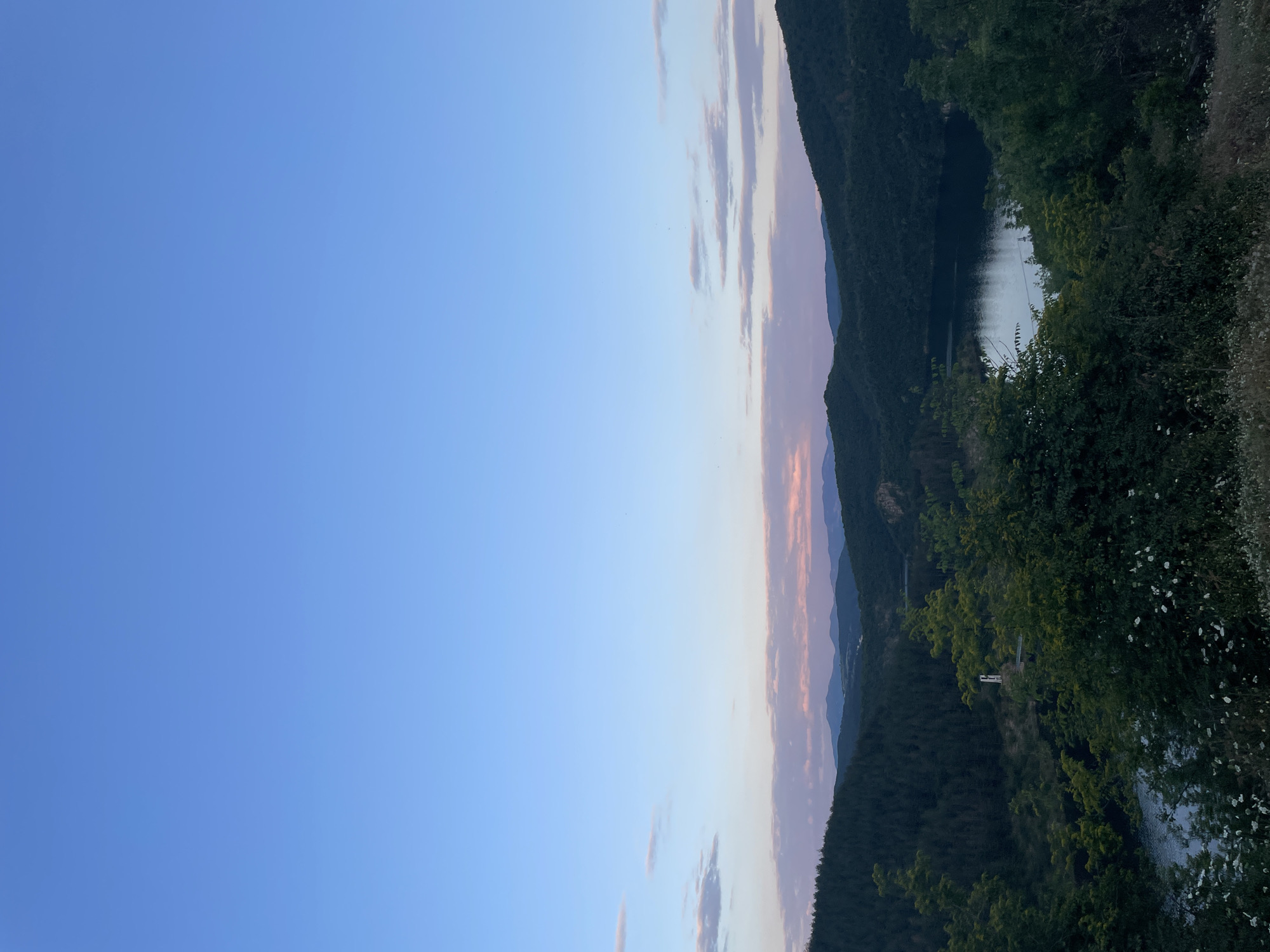 This image depicts a serene landscape of rolling hills and a calm body of water, possibly a lake or reservoir, at dusk. The sky transitions from a pale blue at the top to soft hues of pink and purple near the horizon, indicating the end of the day. Lush green trees and dense forest cover the slopes of the hills. In the mid-ground, a white structure, possibly a monument or a building, is visible on a slight incline. The foreground features wild vegetation, including grasses and small white flowers. The overall impression is one of tranquility and natural beauty. The location is the city of Suhovo, Bulgaria.