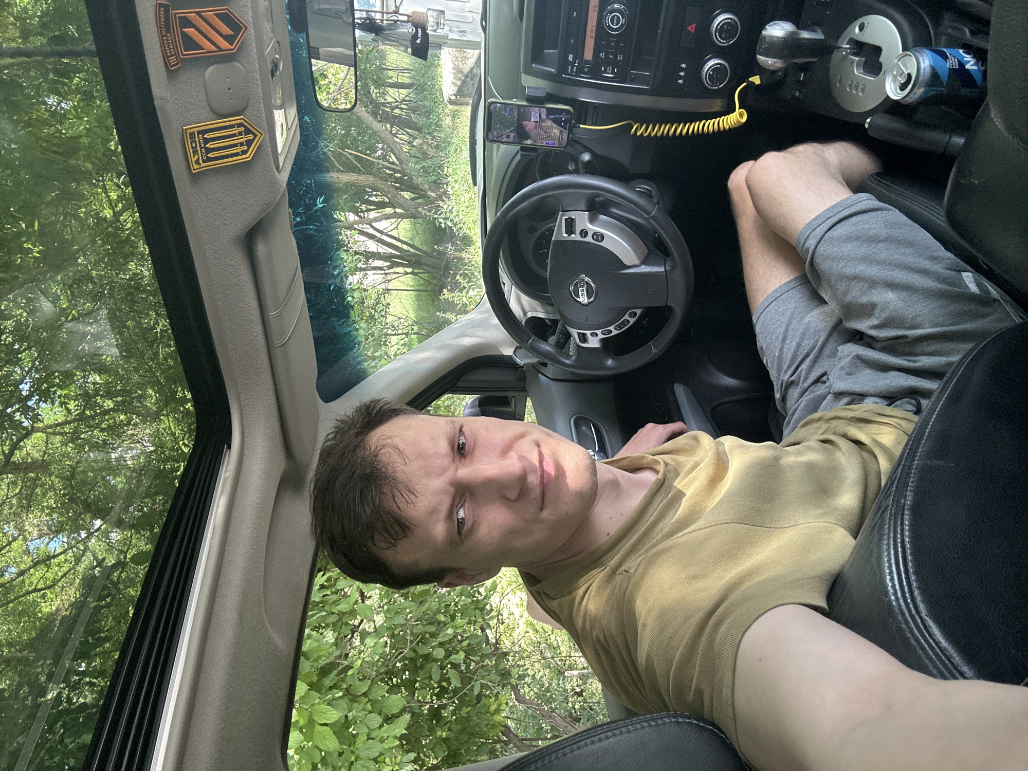 The media file is a selfie taken from inside a car. The subject is a man with short brown hair and light skin, wearing a yellow t-shirt and grey shorts. He is looking at the camera with a slight smile.
The setting appears to be outdoors, with lush green trees and foliage visible through the car's windows and sunroof. Sunlight filters through the leaves, suggesting it is daytime and the weather is likely pleasant.
Inside the car, the dashboard and steering wheel are visible. A smartphone is mounted on the dashboard, displaying a video call. A yellow charging cable is connected to the phone. A can of what appears to be "Noni" juice is placed in the cup holder. There are also two patches affixed to the car's interior trim, one featuring orange and black diagonal stripes, and another with three yellow swords on a black background.
The man's relaxed posture and the casual attire suggest a leisurely activity, possibly a break during a road trip or simply enjoying the outdoors from the comfort of his car. The location context provided indicates this scene is in Makiv, Ukraine.