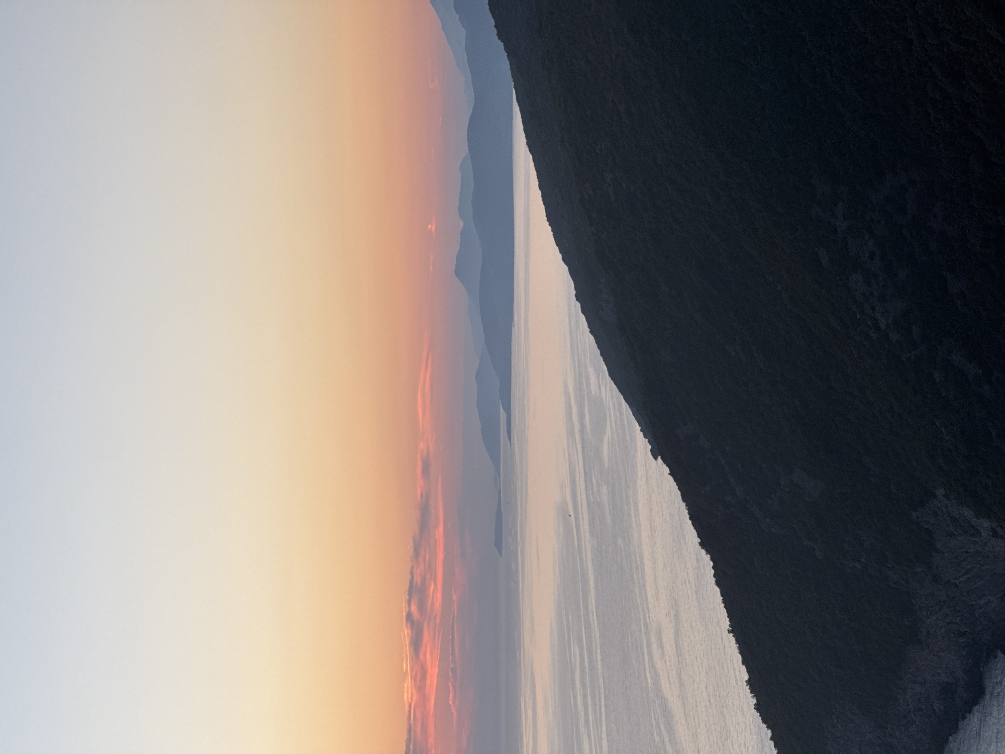 The image depicts a coastal landscape at dusk. A dark, tree-covered mountainside dominates the foreground, sloping down towards a calm sea. The sea reflects the soft hues of the sky, transitioning from a pale blue at the top to warm oranges and pinks near the horizon. In the distance, a series of silhouetted islands or peninsulas are visible against the fading light. The scene is serene and unpopulated, evoking a sense of natural tranquility. The specific location in Zagora, Montenegro is implied by the context provided.