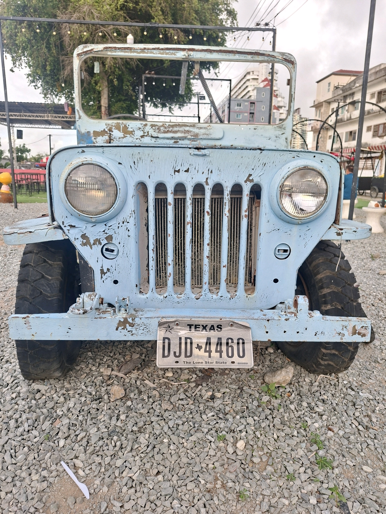 An aged, light blue Jeep is parked on a gravel lot in Pattaya City, Thailand. The Jeep's paint is heavily chipped and weathered, revealing rust underneath. It has a vertical grille with seven slats and two round headlights. A Texas license plate reading "DJD 4460" is affixed to the front bumper. The Jeep is surrounded by small, gray stones. In the background, trees, buildings, and some outdoor seating areas with red tables and chairs are visible. The sky is overcast. A single white plastic utensil lies on the ground in the foreground.