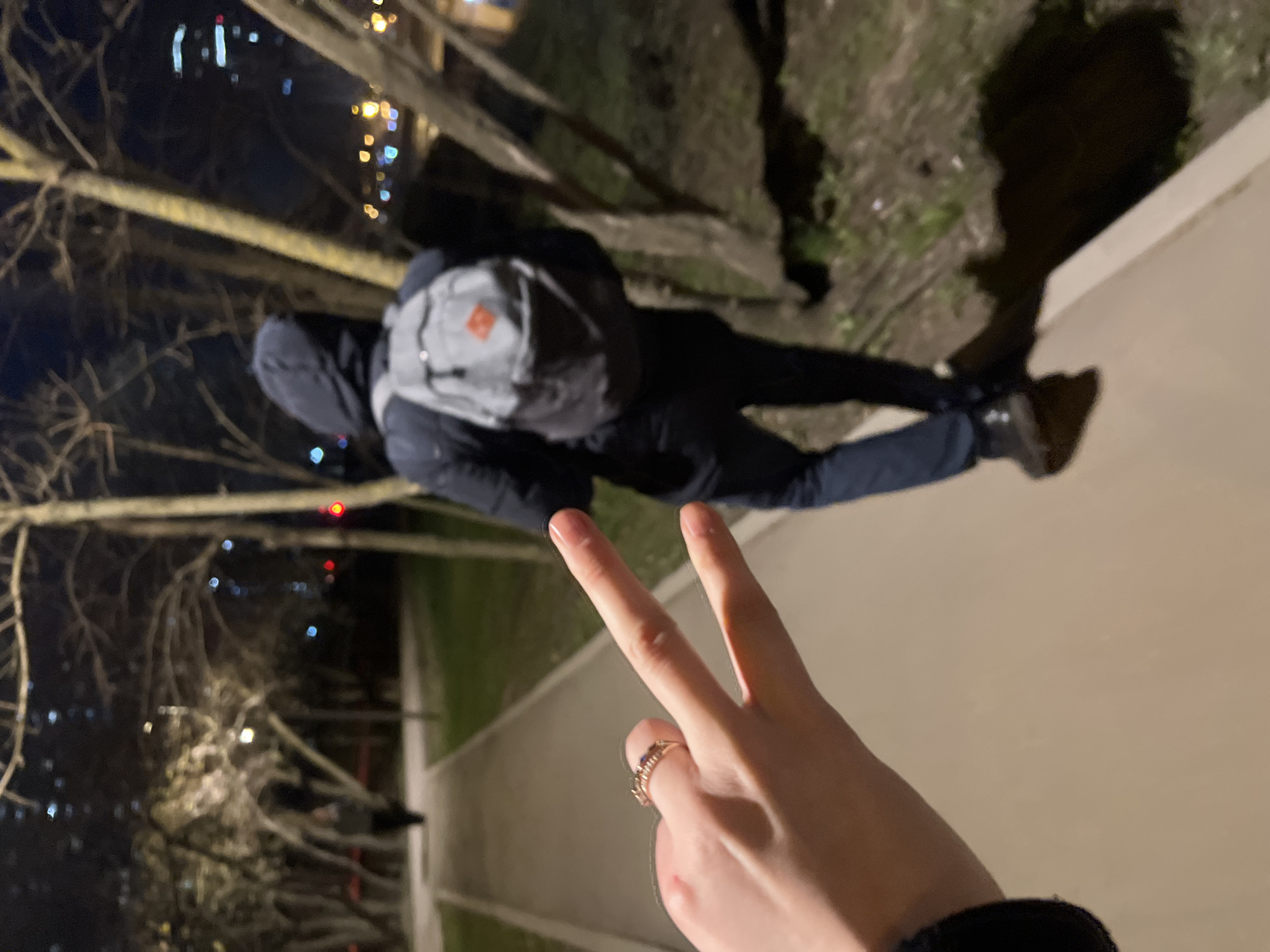 The image shows a person lying on a sidewalk in Khimki, Russia. The person is wearing a hooded jacket, dark pants, and a backpack. Their face is not visible. Another person's hand is in the foreground, making a peace sign. In the background, there are bare trees and city lights, suggesting it is nighttime. A sidewalk is visible, with some greenery adjacent to it.