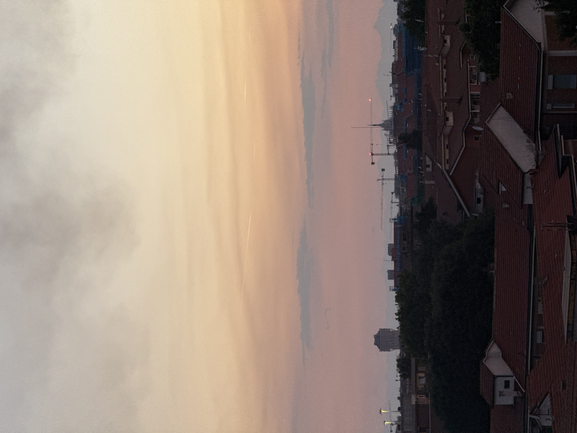 The image depicts a cityscape at sunset, likely from a high vantage point, overlooking a dense urban environment in Milan, Italy. The sky transitions from a pale yellow at the horizon to a soft pink and then to a muted gray, with wispy clouds and a faint contrail visible. Below the sky, the skyline is dominated by a mosaic of red-tiled rooftops of residential buildings.
In the middle ground, several prominent structures rise above the rooftops. A distinct, cylindrical building with a lattice-like facade stands out, possibly a water tower or a unique architectural landmark. Further in the distance, a cluster of cranes suggests ongoing construction or development within the city. A tall, spired building, likely a cathedral or church, can be seen among the cranes, a common feature of Milan's historical architecture. Beyond the city, hazy, silhouetted mountains form a distant backdrop.
The foreground is occupied by more red-tiled roofs, some with visible chimneys and dormer windows. The lighting suggests it is late afternoon or early evening, with the warm tones of the sunset casting a soft glow over the scene. No people are discernible in the image, and the focus is entirely on the urban landscape and the atmospheric sky. The overall impression is one of a sprawling city bathed in the serene light of dusk.