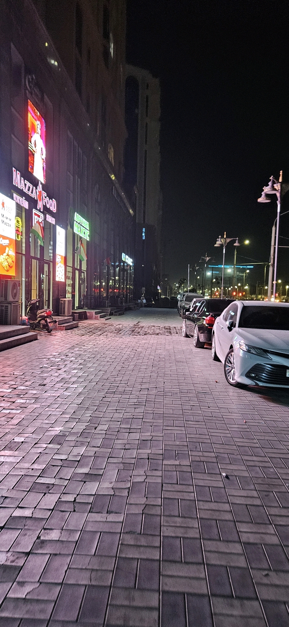 The image depicts a nighttime scene on a wide, paved pedestrian walkway lined with buildings on the left and parked cars on the right. The walkway is paved with rectangular bricks, some of which appear cracked or uneven. The buildings on the left are illuminated by numerous neon signs, including one for "MAZZA FOOD" which features a large, vibrant advertisement. Several air conditioning units are visible on the lower levels of the building. A scooter is parked on the walkway near the entrance.
On the right side of the walkway, several cars are parked in a line. The foreground shows the front passenger side of a white sedan, likely a Toyota Camry, with its wheel clearly visible. Further down the street, other vehicles, including a dark-colored sedan, are parked. Streetlights with ornate lamps illuminate the area, casting long shadows on the walkway. In the background, more buildings and city lights are visible, suggesting a commercial or urban environment. The overall impression is of a well-lit commercial street at night.
This scene is located on Rudaki Avenue in Dushanbe, Tajikistan, specifically in the vicinity of the Mazza Food establishment. Rudaki Avenue is a major thoroughfare in Dushanbe, known for its commercial and administrative buildings, as well as its vibrant nightlife.