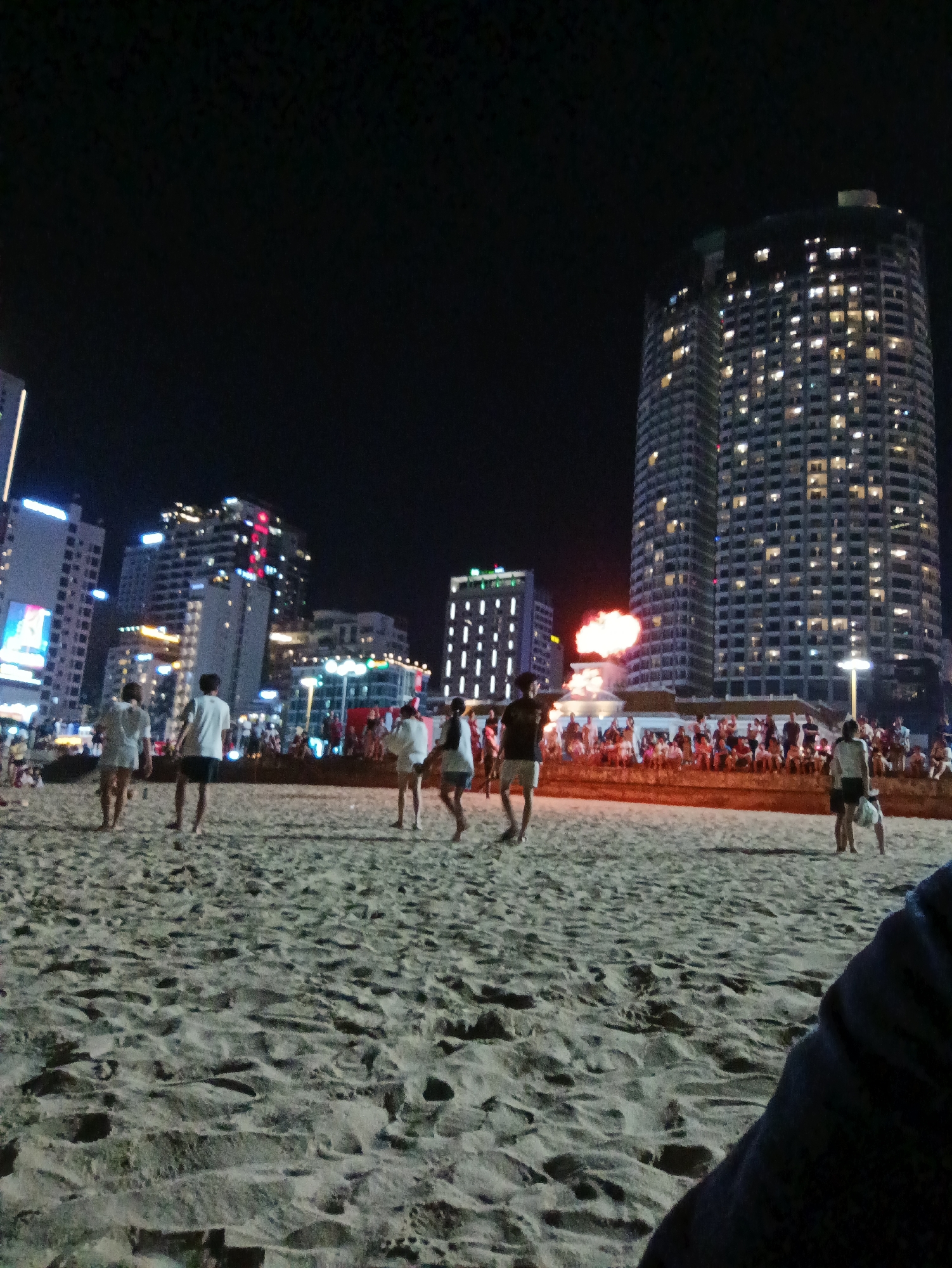 The image depicts a nighttime scene on a sandy beach in Nha Trang, Vietnam, with a backdrop of illuminated city skyscrapers. In the foreground, several people are walking on the beach. Further back, a crowd is gathered, watching an event that appears to involve fire, possibly a performance or fireworks. Tall buildings with numerous lit windows dominate the skyline, indicating a densely populated urban area. The sand is visible with footprints and slight undulations. The overall atmosphere suggests a lively evening in a coastal city.