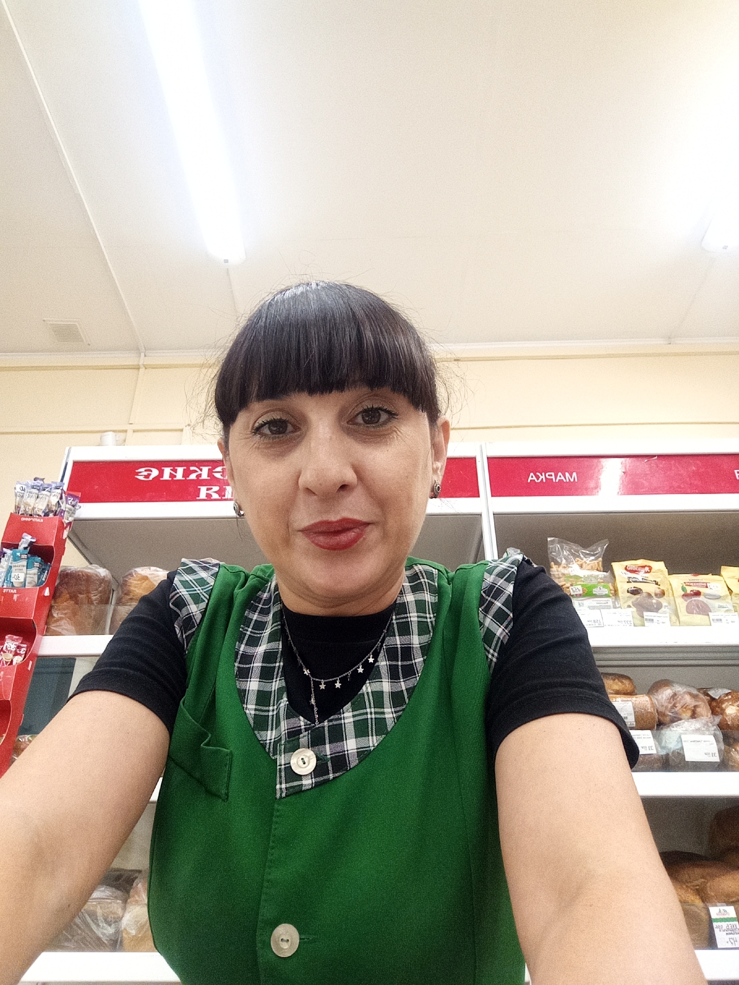A medium shot from a selfie perspective shows a woman with dark hair and bangs, wearing a green vest over a black shirt. The vest has a plaid collar and two white buttons. She is wearing a necklace with star charms. Behind her, shelves are stocked with various food items, including bread and packaged goods. Red signage with white lettering is visible on the shelves, indicating "РЫБА" (fish) and "МАРКА" (brand). The lighting suggests an indoor commercial environment, possibly a grocery store, with overhead fluorescent lights. The overall scene is well-lit and appears to be a casual snapshot.