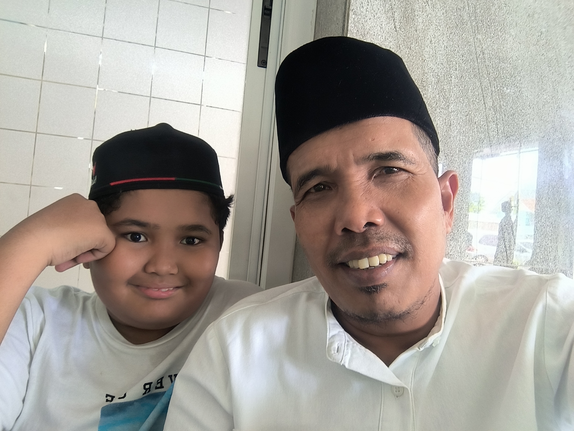 The image shows two people, likely a father and son, posing for a selfie. The father is on the right, wearing a white shirt and a black peci hat. He is smiling at the camera. The son is on the left, wearing a white t-shirt and a peci with red and green trim. He is resting his head on his hand and smiling.
The background behind them appears to be a tiled wall on the left and a frosted glass window on the right. The reflection in the glass reveals an outdoor area with light-colored vehicles parked, and a building is visible. Based on the attire and appearance of the individuals, and the information that they are likely in Lhokseumawe, Indonesia, this picture may have been taken to celebrate Eid al-Fitr.