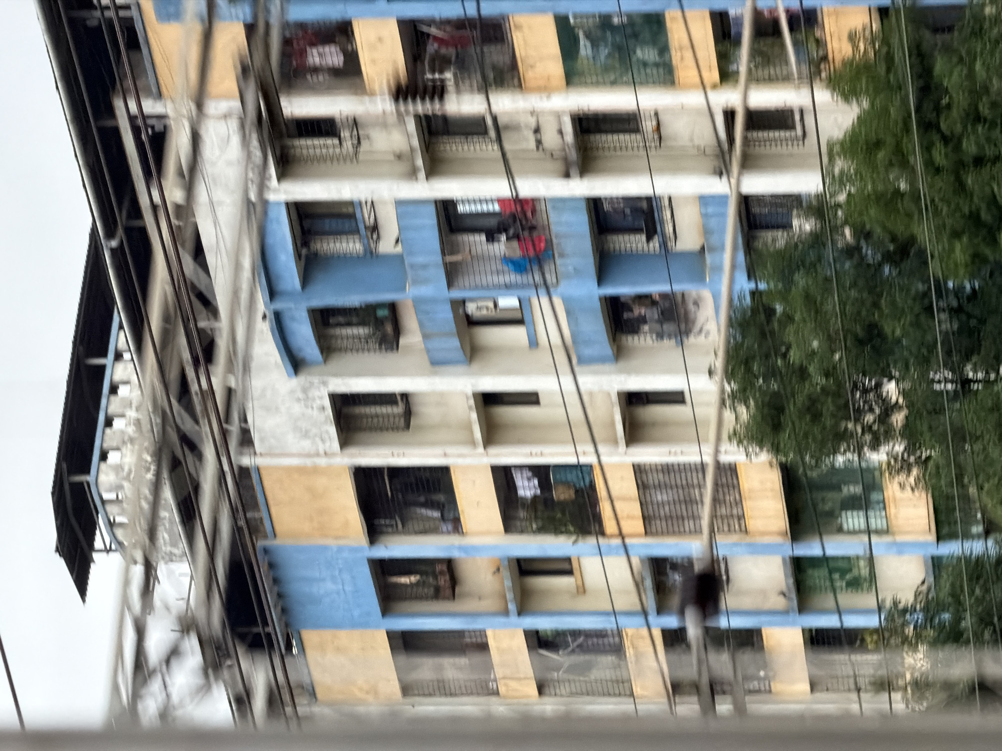 The image shows a multi-story apartment building in Vasai-Virar, India. The building has a blue and yellow facade with several windows and balconies. Electrical wires are visible in the foreground, obscuring parts of the building. Trees are growing in front of the building. The sky is overcast. There are no visible people in the image, but some laundry is hanging on balconies. The architectural style appears to be typical of residential buildings in urban India.