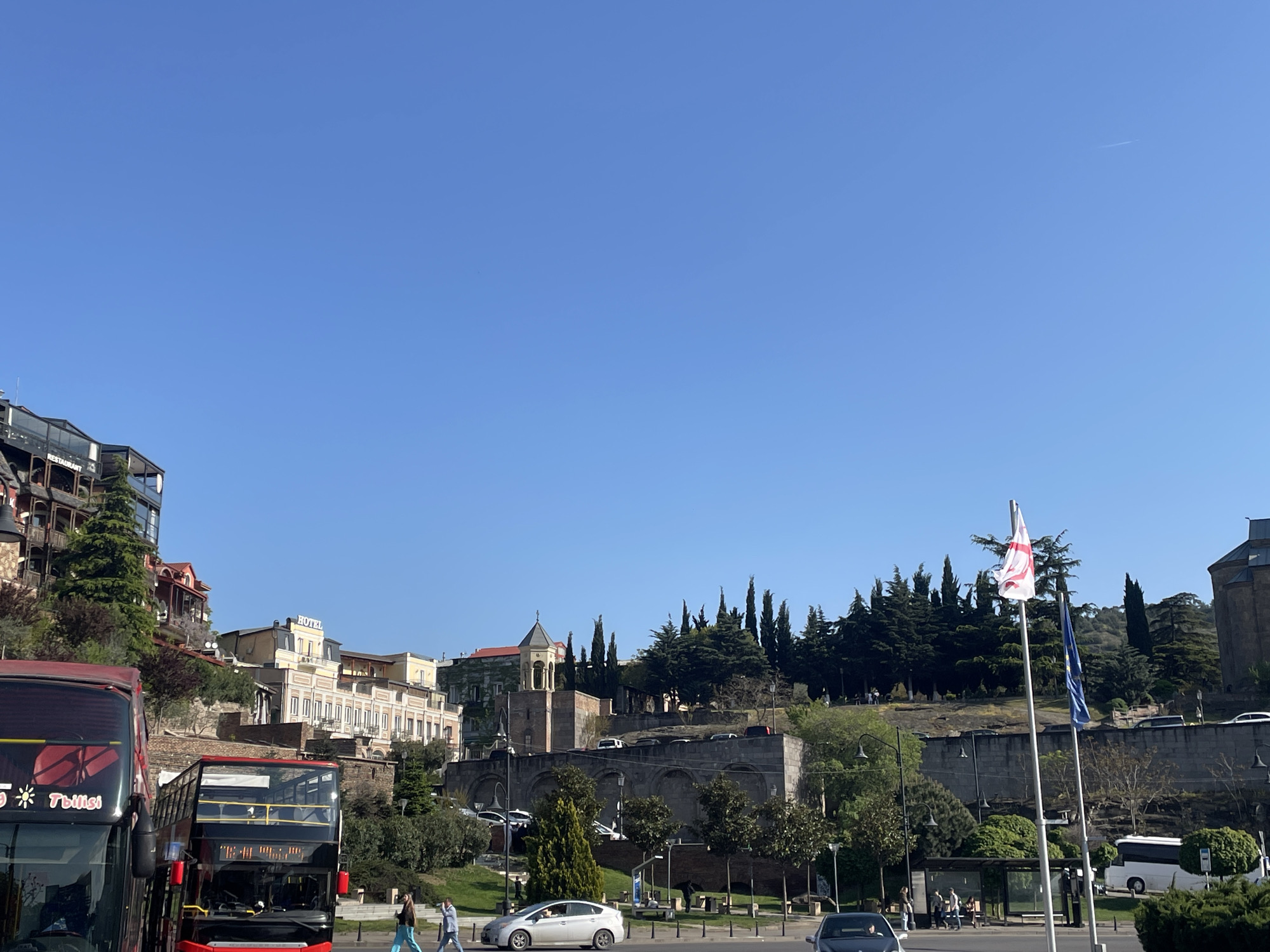 This daytime image captures a vibrant street scene in Tbilisi, Georgia, under a clear, bright blue sky. In the foreground, two red double-decker tour buses are prominent, with the one on the left displaying "Tbilisi" and partially visible "Polski" text. Several pedestrians are visible walking along sidewalks, and various cars, including a white Toyota Prius, are on the road. To the right, two flagpoles display the Georgian national flag (a red cross on a white field with four smaller crosses) and the European Union flag.
The middle ground features a green, landscaped area with numerous trees, notably tall, slender cypress trees. A large, ancient-looking stone wall with arched openings supports an elevated section, providing a base for more greenery and buildings.
In the background, on a hillside, a mix of architectural styles is apparent. On the left, modern multi-story buildings with balconies and glass facades are visible, one featuring a "HOTEL" sign and another a "RESTAURANT" sign. Towards the right, a more traditional building with a distinctive bell tower structure stands out against the sky. The scene depicts daily urban activity, with people walking and vehicles moving, set against a backdrop of historic and contemporary architecture typical of Tbilisi.