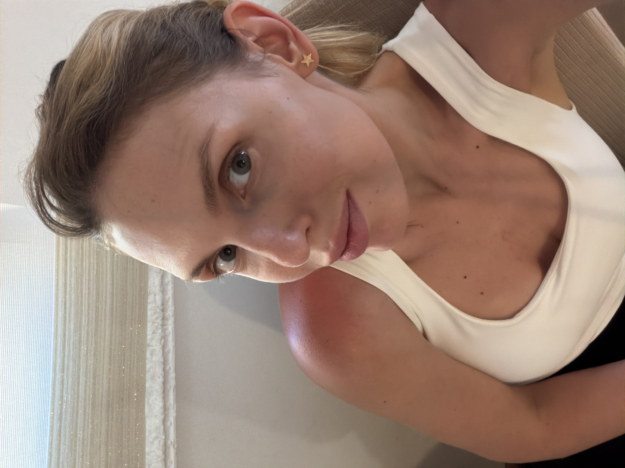 A woman is seen at close range in an indoor setting. She has light skin, blonde-brown hair, and wears a white tank top. A gold star earring is visible in her left ear. Her hair appears styled, and she looks directly towards the camera with a slight smile. The background includes a window with light-colored curtains and a portion of a beige-colored surface. There are no immediate visual cues indicating the location is Budva, Montenegro.