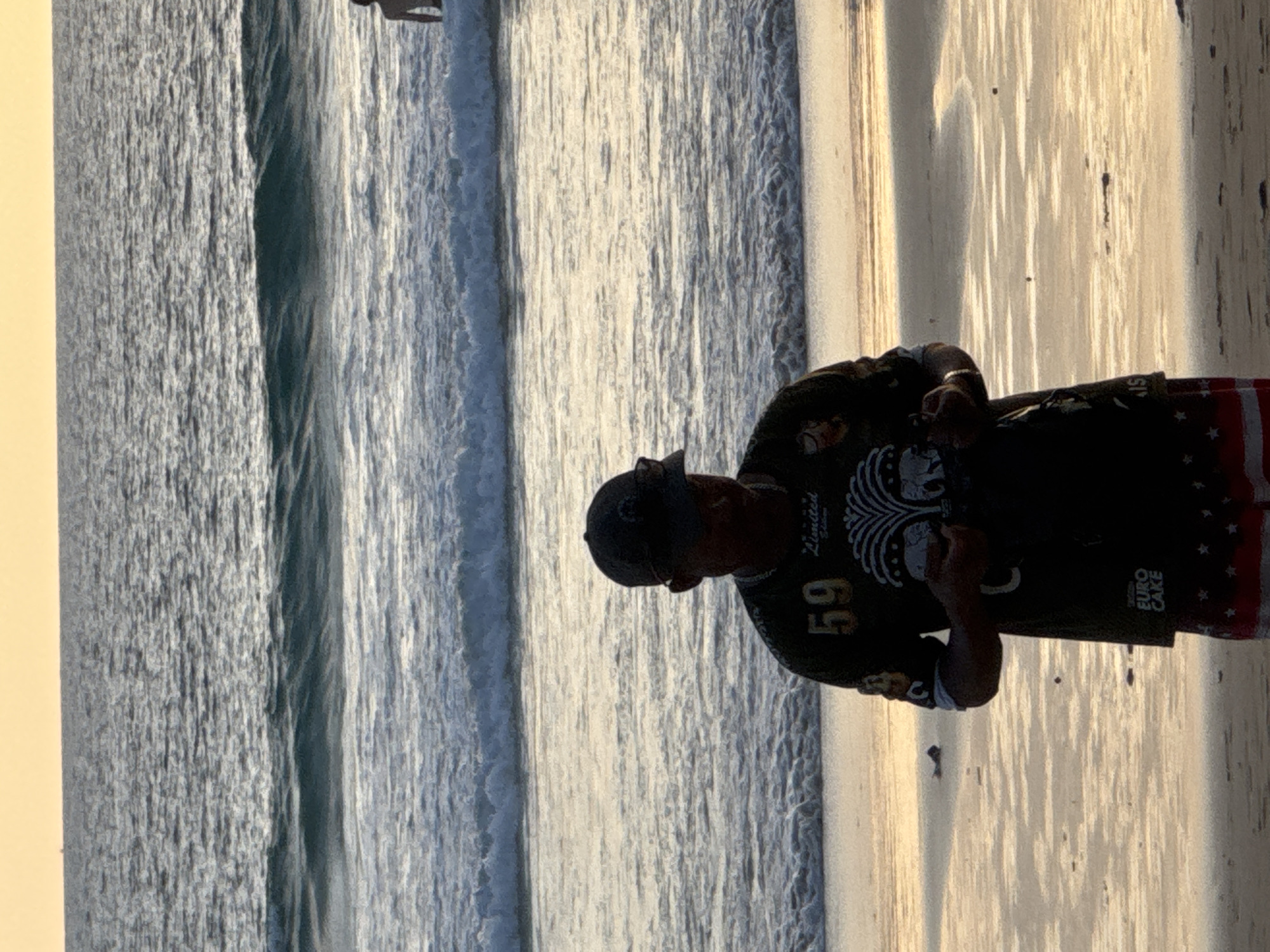 An adult male wearing a baseball cap, a dark t-shirt with graphics of elephants and the number 59, and patterned shorts stands on a beach at Karon, Thailand. He is holding a dark object in his hands. The ocean with gentle waves stretches behind him towards the horizon, and the sky above is a soft yellow-orange, indicative of sunrise or sunset. The sand on the beach is wet in places, with small pools of water reflecting the sky. Another person, a child, is partially visible in the water in the distance to the right.