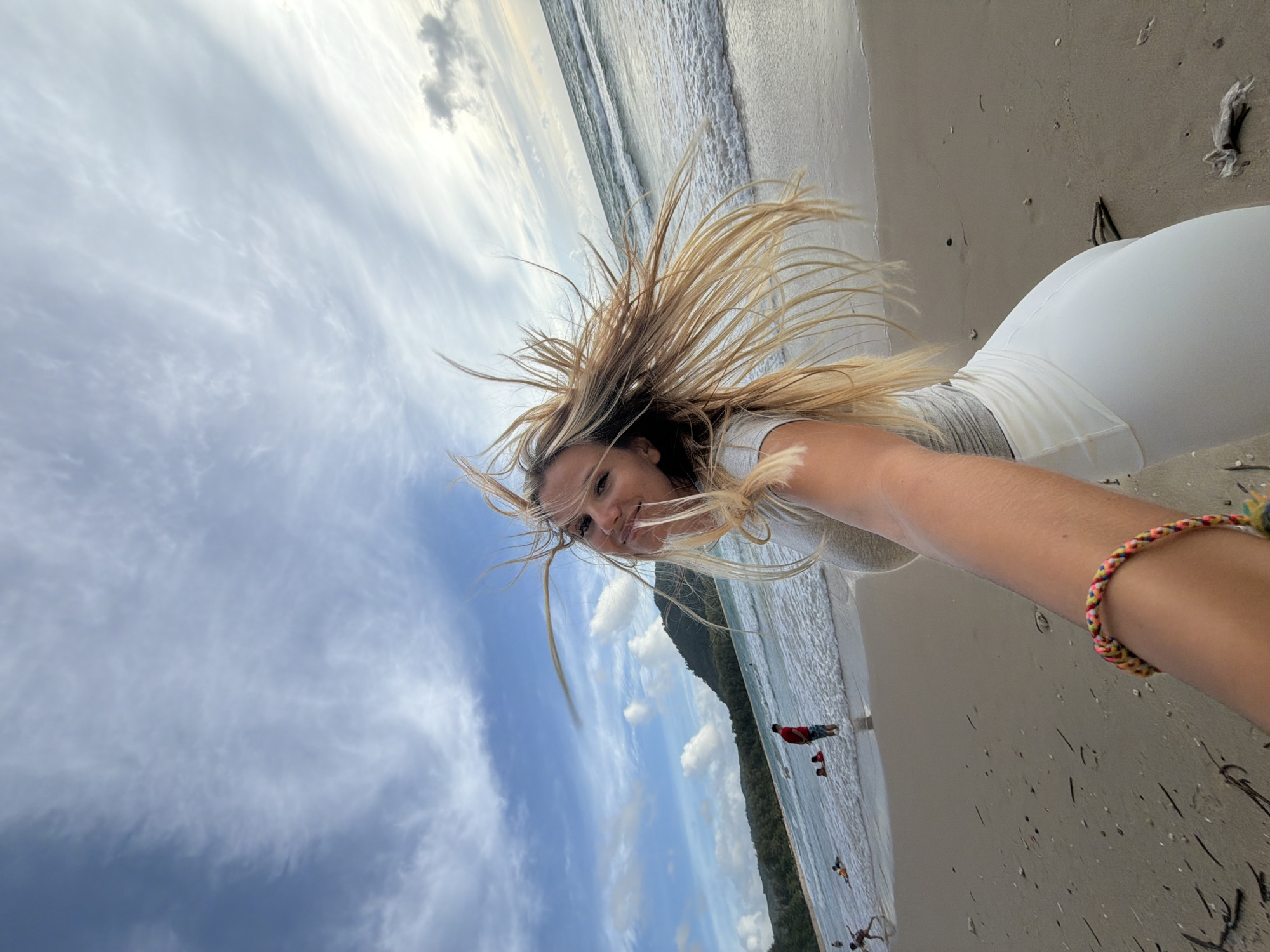 This is a selfie taken on the beach in Sakhu, Thailand. A woman with long, blonde hair that is being blown by the wind is in the foreground, looking back at the camera and smiling. She is wearing a light-colored top and white pants. On her left wrist, she has a braided bracelet with multiple colors. In the background, the ocean meets the sandy shore. There are several people in the water and on the beach, including a man in a red shirt. Lush, green hills rise behind the beach. The sky is partly cloudy with a mix of blue and white.